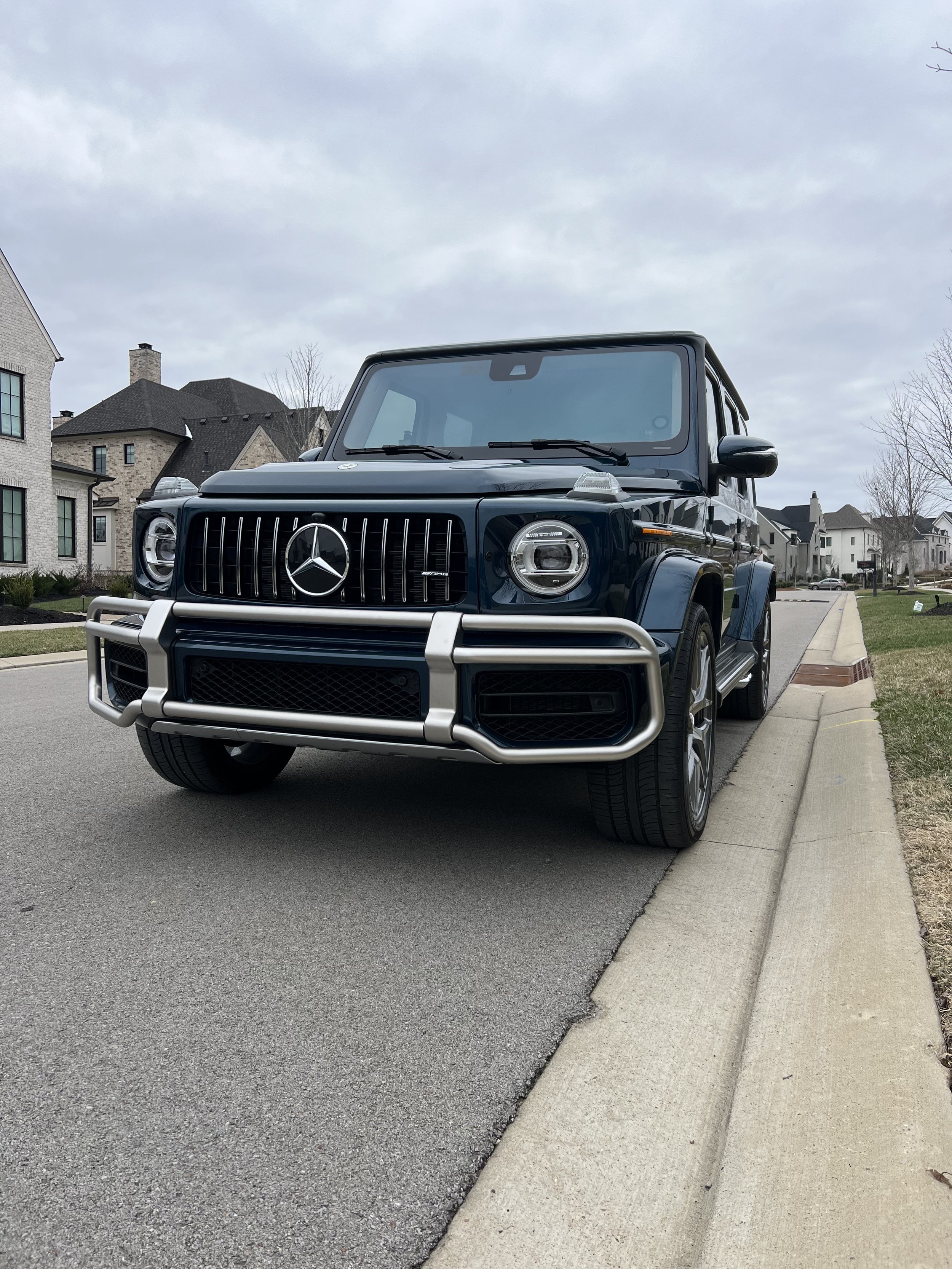 A black Mercedes-Benz G-Class SUV parked on a residential street with houses and trees in the background.