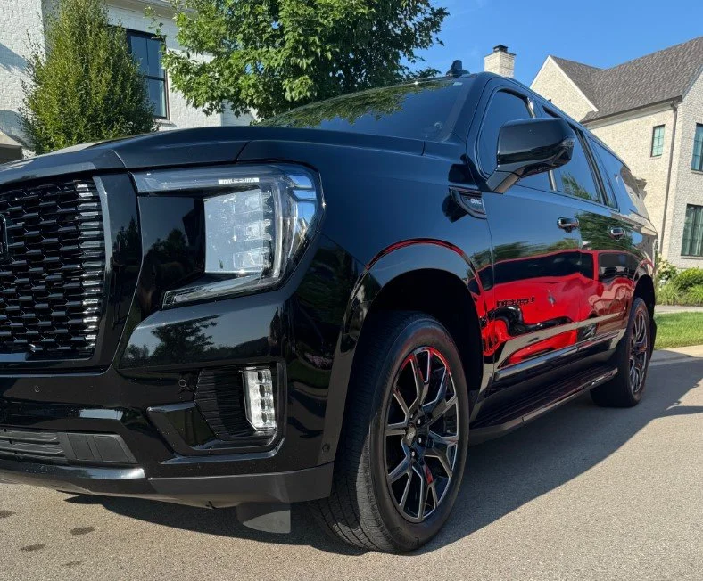 Black SUV parked on the street in front of residential houses with trees and blue sky in the background.