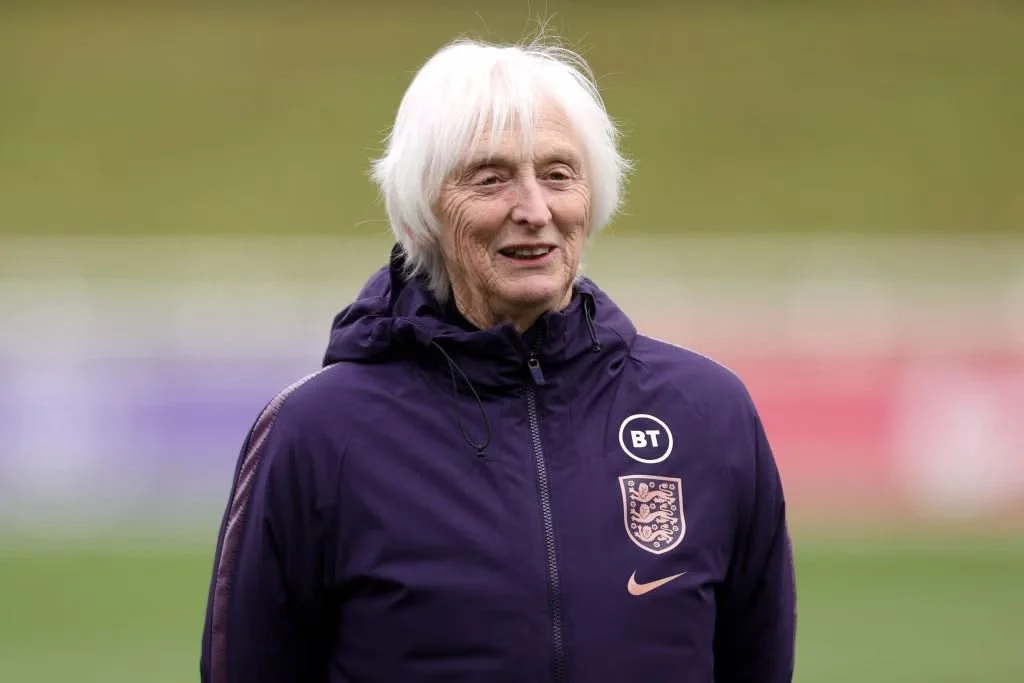 An elderly woman with white hair smiling outdoors, wearing a purple Nike sports jacket with the England football team emblem and sponsor logos.