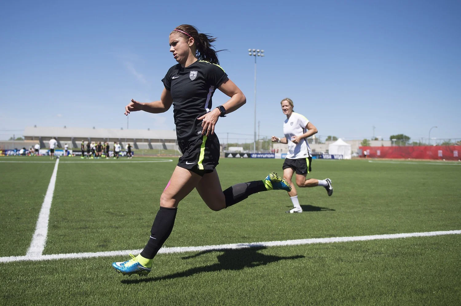 Two female soccer players running on a green soccer field during practice or game under a clear blue sky.