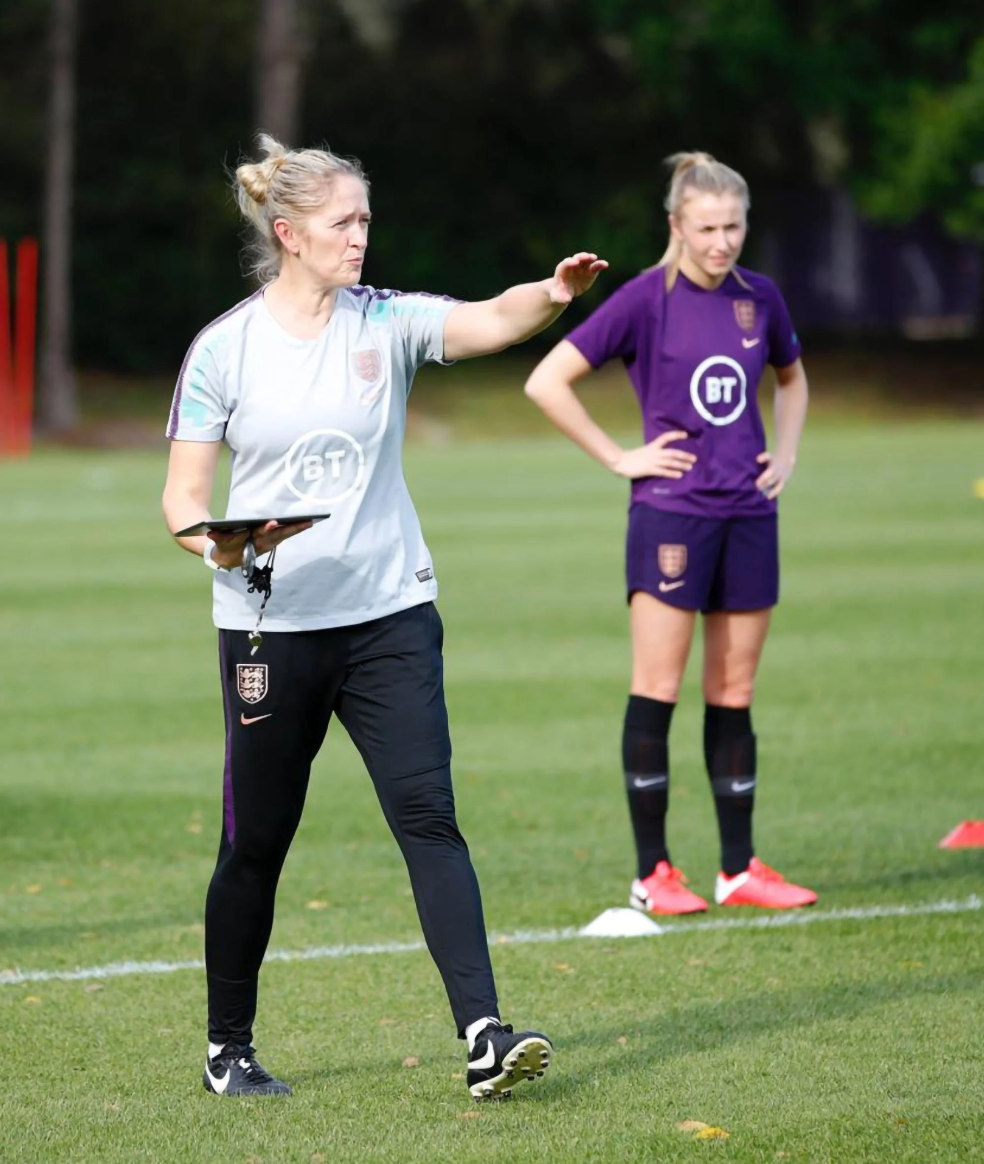 A woman soccer coach giving instructions to a female soccer player during practice on a grassy field, with the player standing behind, listening with her hands on her hips.