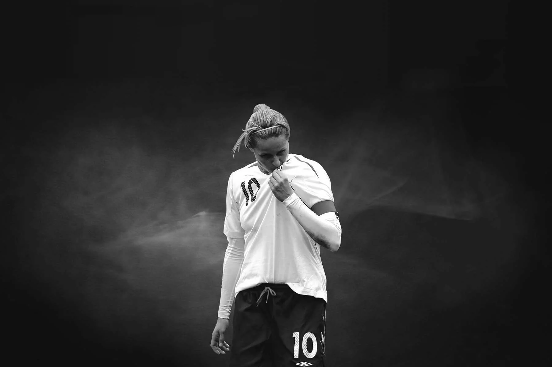 Black and white photo of a female soccer player in a jersey with the number 10, shirt lifted to reveal her face, standing against a dark background.