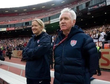 Two people standing at a sports stadium, wearing USA jackets, with a crowd in the background.