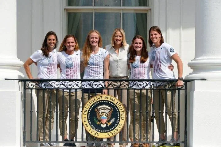 Six women standing on a balcony in front of a window, with the seal of the President of the United States hanging from the railing.