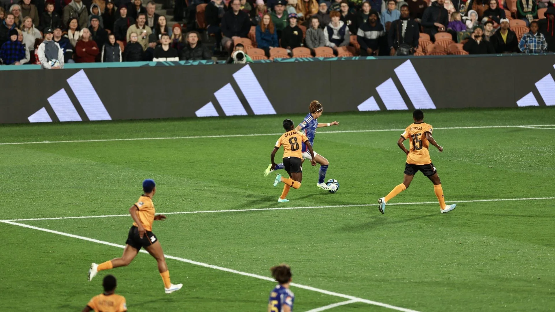 A soccer player in a blue uniform is kicking a ball while three players in orange uniforms with black shorts and orange socks are nearby on a green field during a match, with spectators watching from the stands.