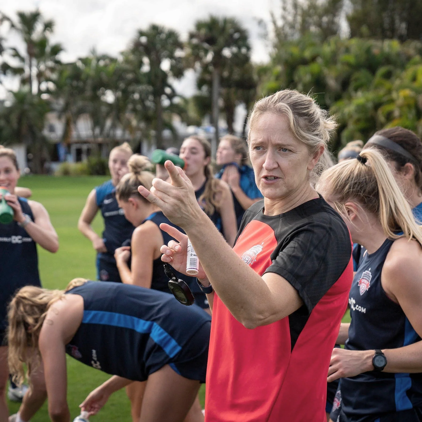 Dawn Scott giving instructions to a group of female athletes on a grassy field during a sports practice or game, with trees in the background.