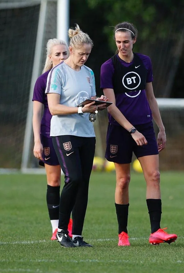 Three women in soccer uniforms standing on a soccer field, looking at a clipboard held by one of them. All wear black knee-high socks, and two have pink soccer cleats. The woman in the middle appears to be coaching or giving instructions, while the other two players listen attentively.