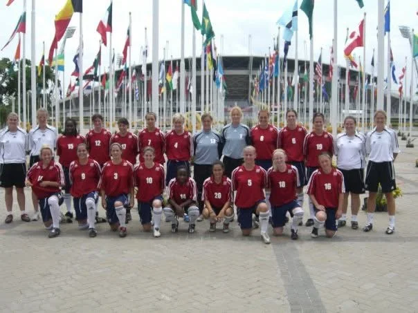 Female soccer team wearing red jerseys and blue shorts, posing for a group photo in front of a display of international flags outside a stadium.