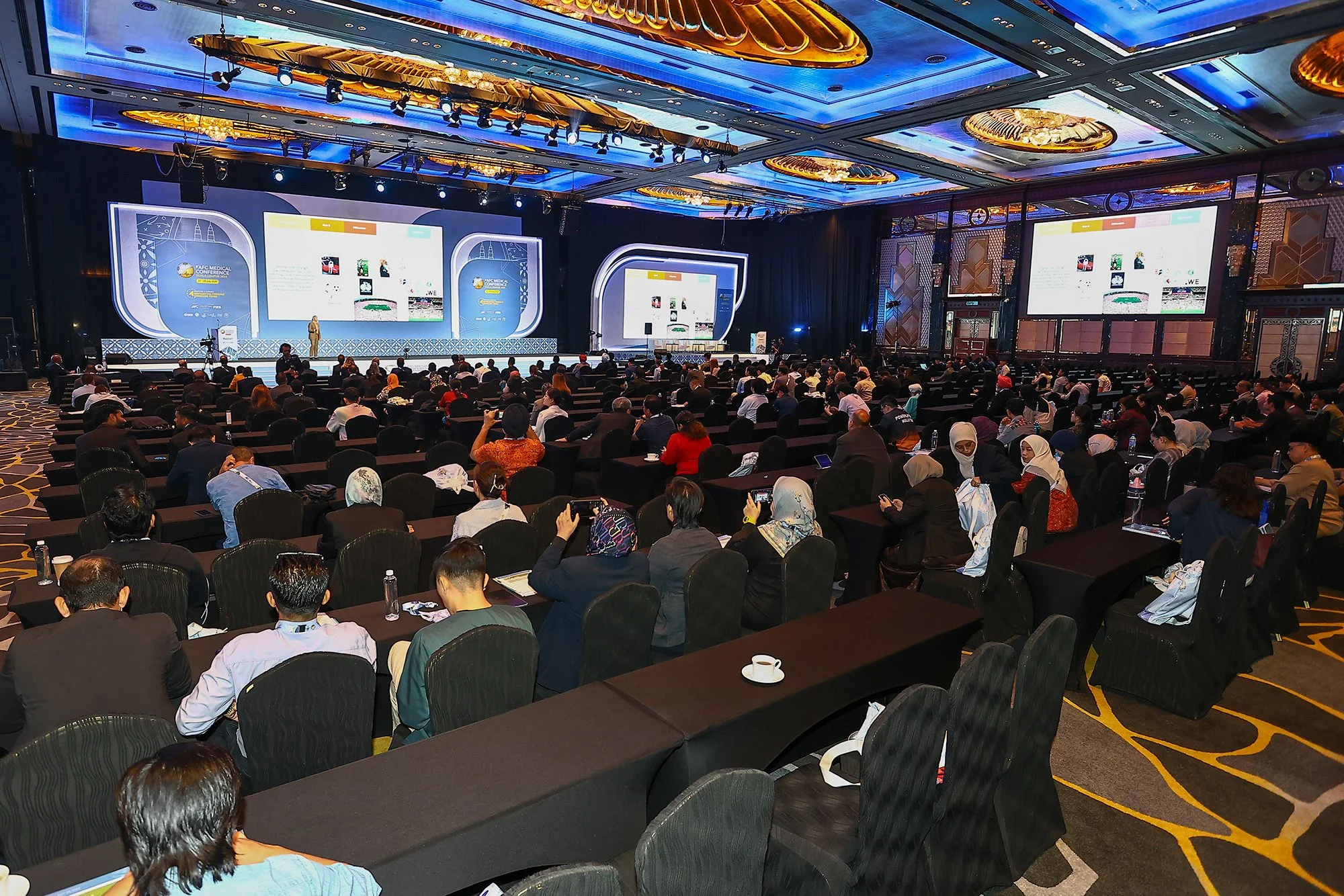 A large conference room filled with seated attendees watching a presentation on a stage with multiple large screens.