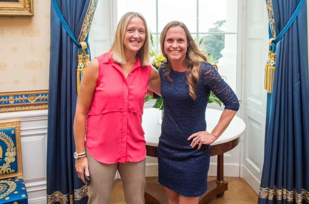 Two women standing together in a luxurious room, smiling, with one wearing a pink sleeveless top and beige pants, and the other wearing a dark blue lace dress. There are blue curtains with gold accents, and a table with a flower arrangement in the background.