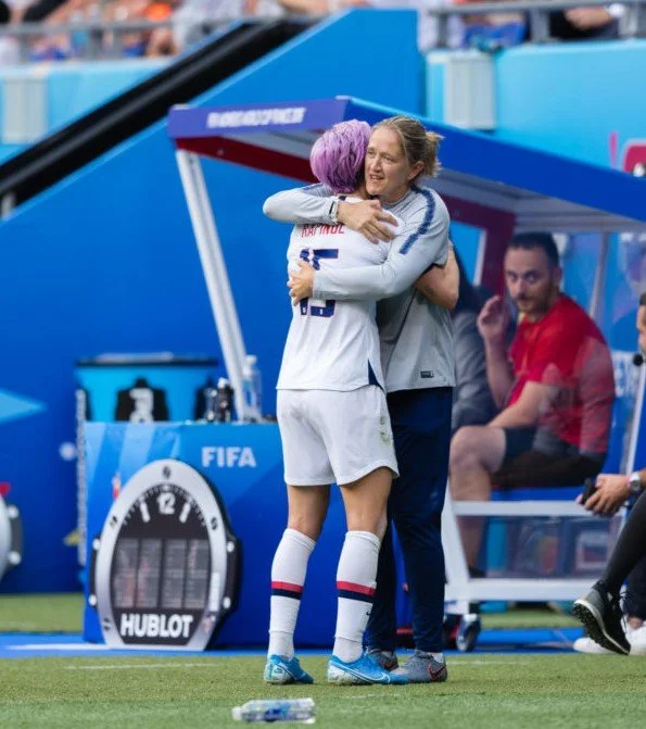 Two female soccer players hugging on the sideline of a soccer field, with one wearing a white uniform and the other with purple hair, smiling and celebrating.