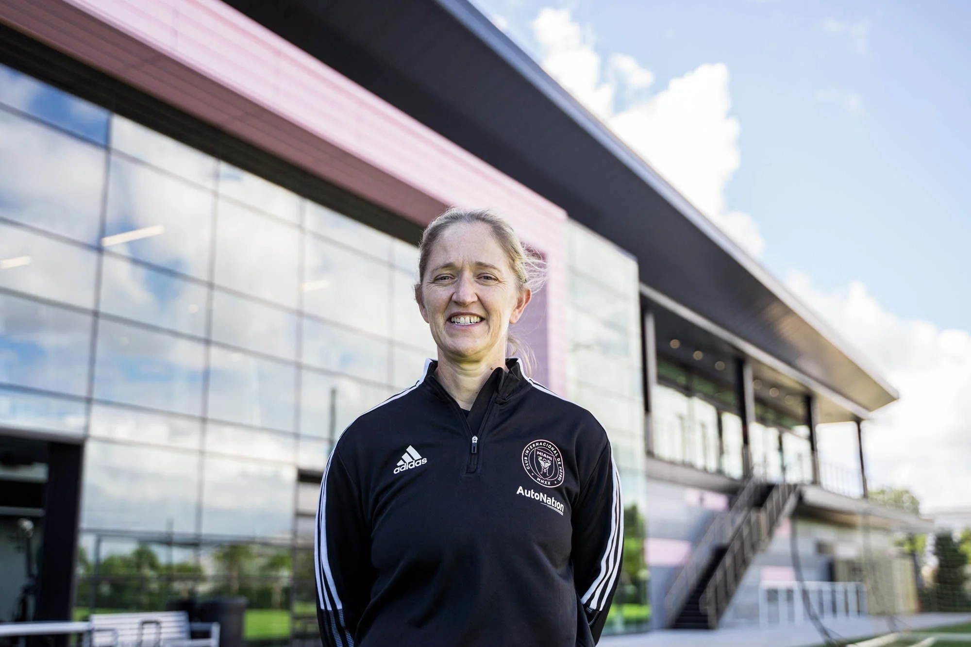A woman with blond hair smiling outdoors in front of a modern glass building, wearing a black Adidas jacket with a sports team logo.