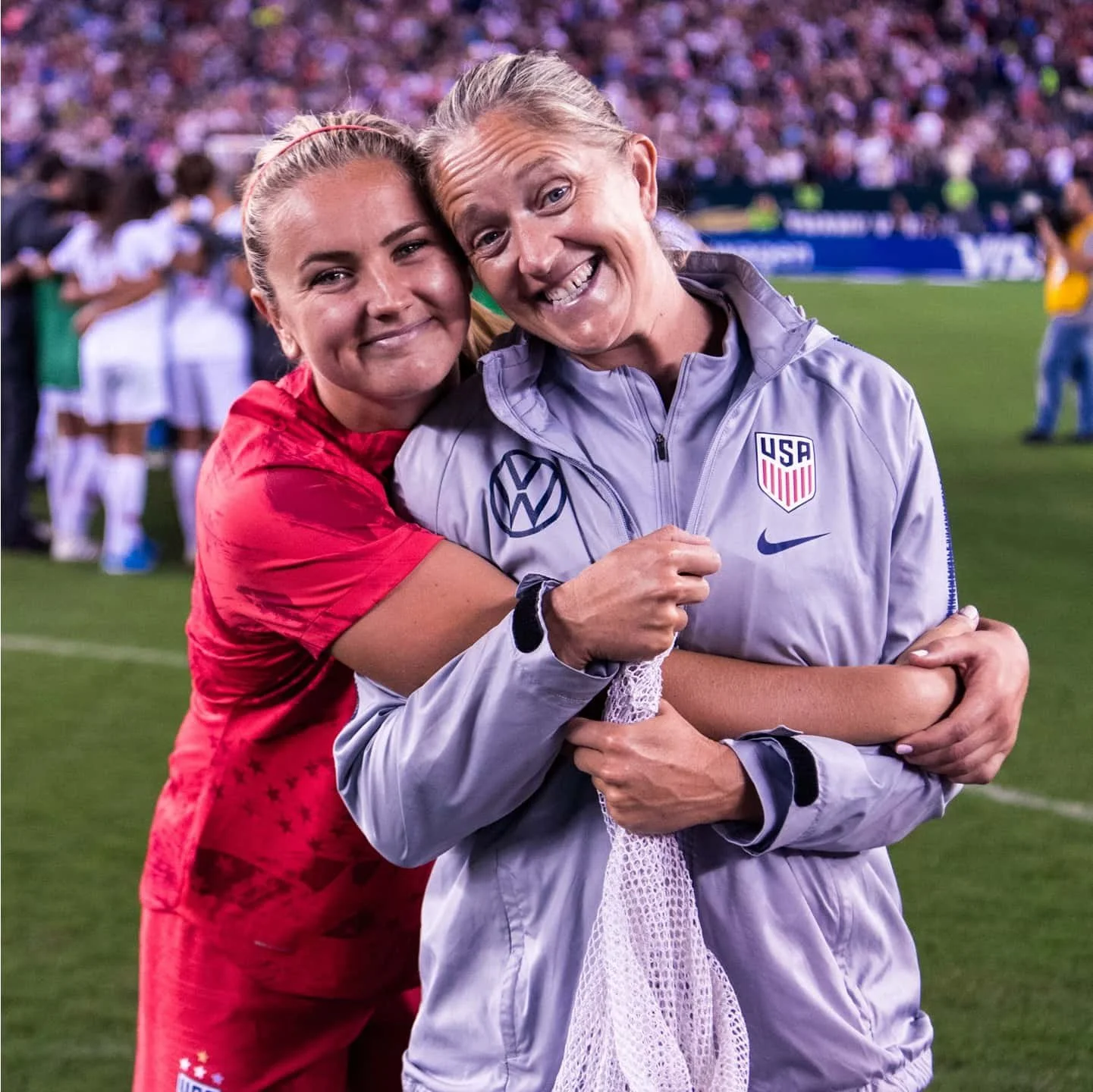 Two women in sports uniforms hugging on a football field, smiling at the camera, with a crowd in the background.