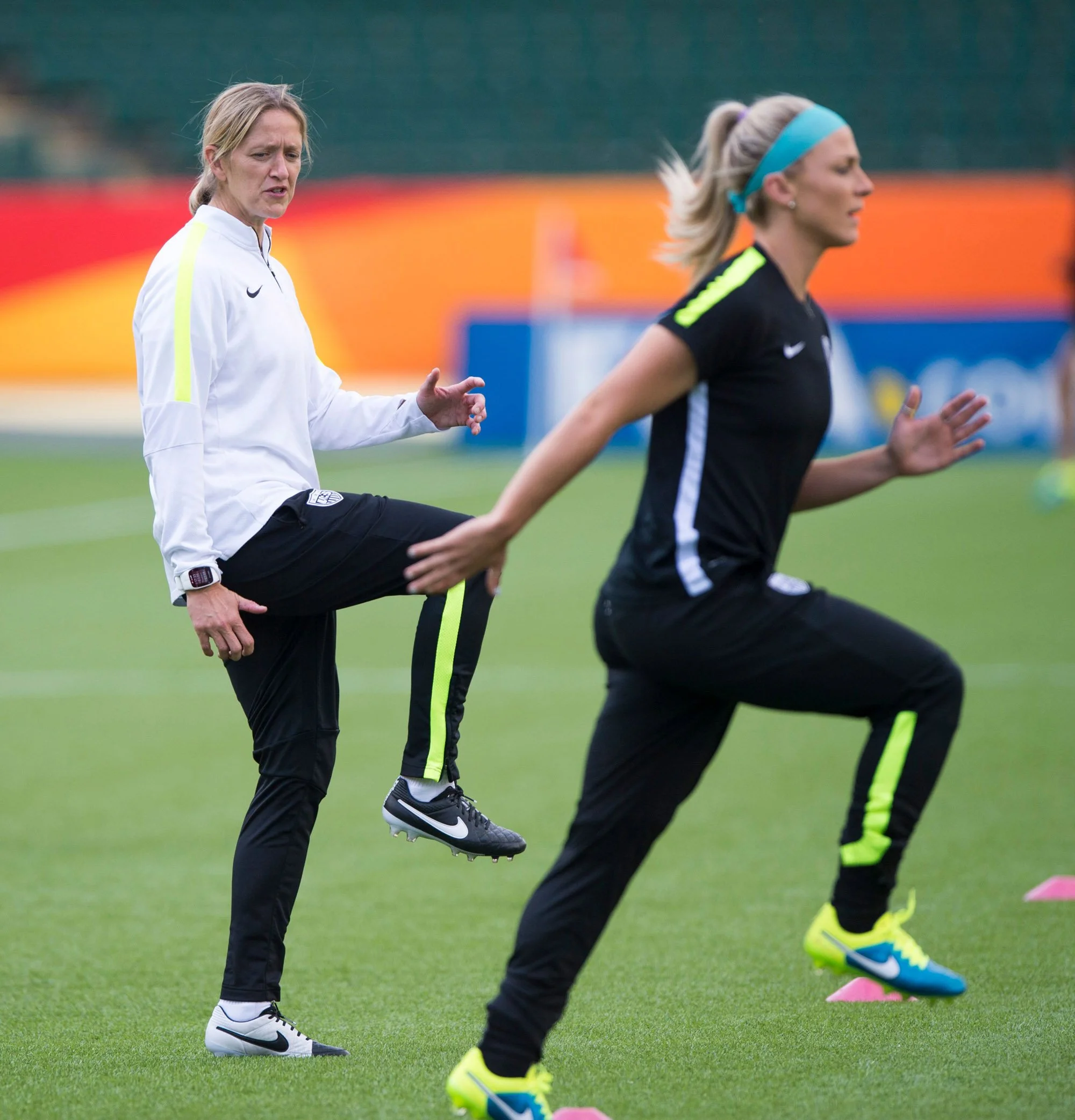 Two women on a soccer field during a training session. One woman, wearing a white jacket and black pants, is stretching her leg while the other, in a black athletic outfit with neon yellow accents and a blue headband, is running times beside pink markers.