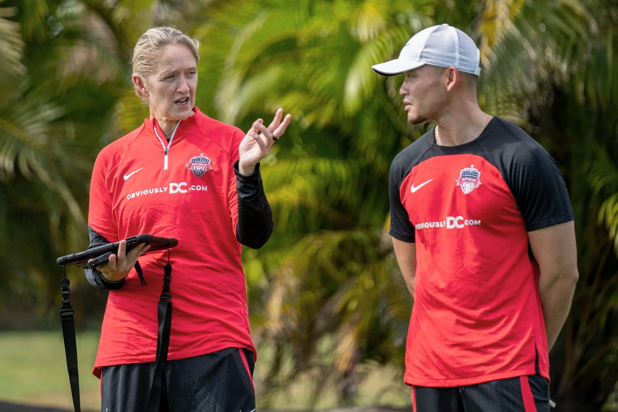 Two soccer players in red and black training jerseys with the Memphis 901 FC logo, having a conversation outdoors with tropical plants in the background.