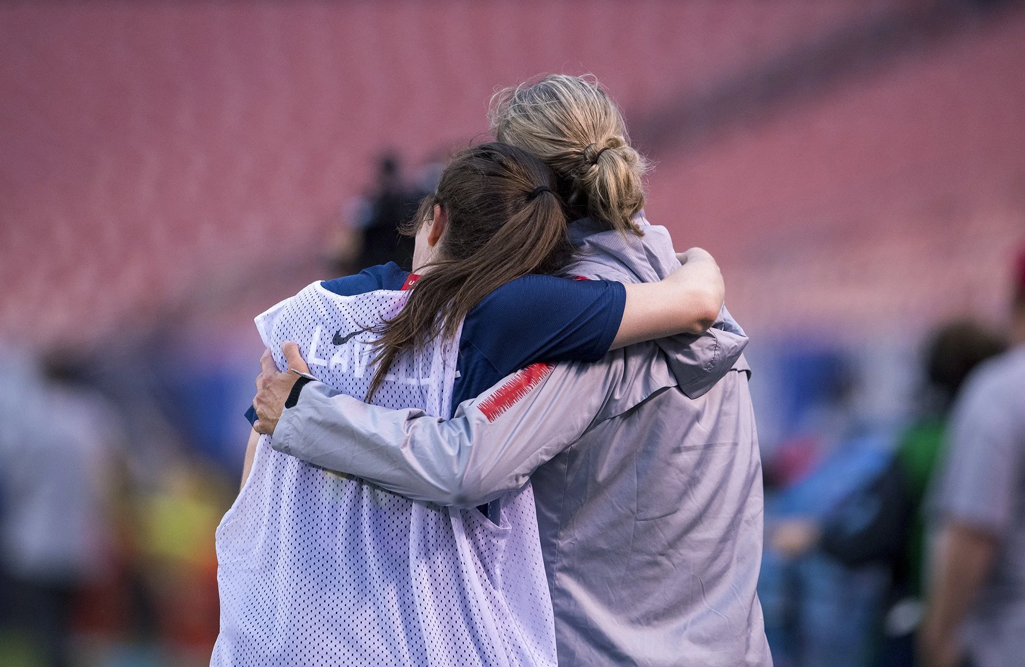 Two women are hugging each other on a sports field, one wearing a sports jersey and the other in a gray jacket, with blurred spectators in the background.