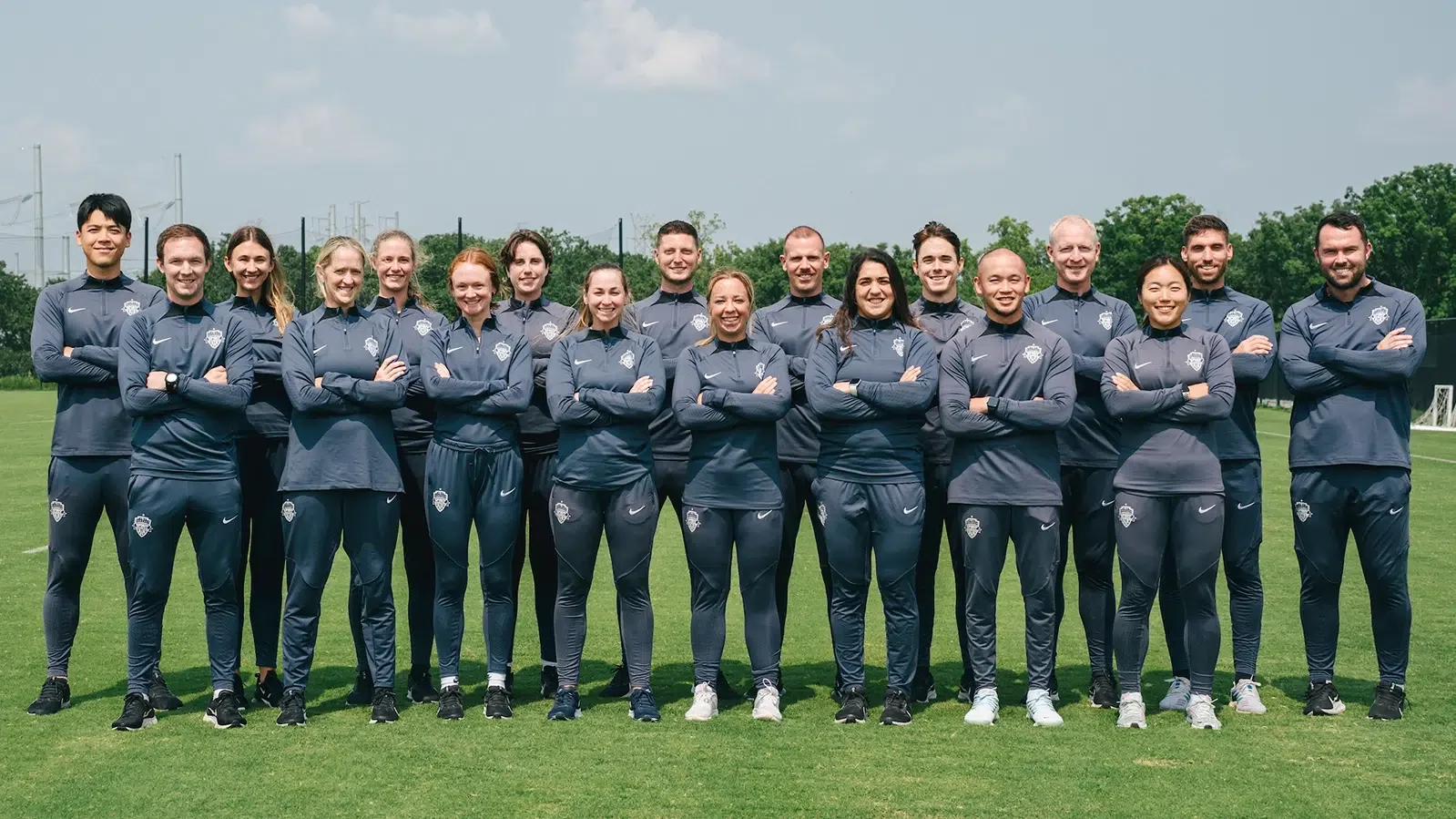 A group of 16 diverse women and men in navy sportswear with a team logo, standing on a soccer field with arms crossed, smiling for a team photo.