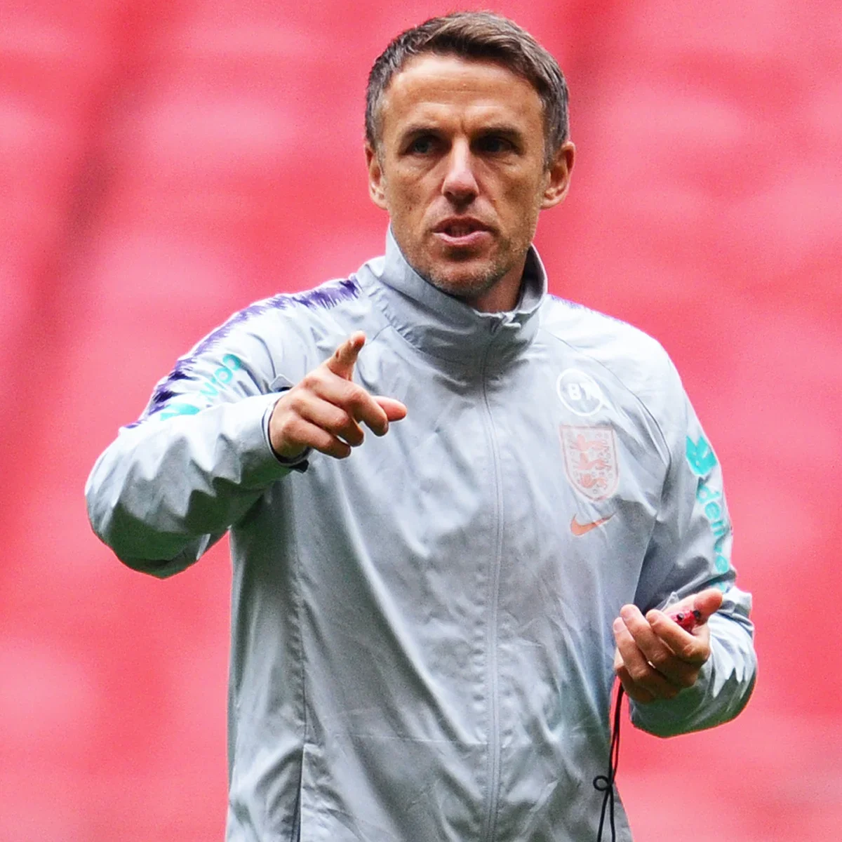 A man with short dark hair and light stubble, wearing a gray sports jacket with the England national football team crest, pointing and holding a small device, possibly a microphone or headphone, with a pinkish-red background.
