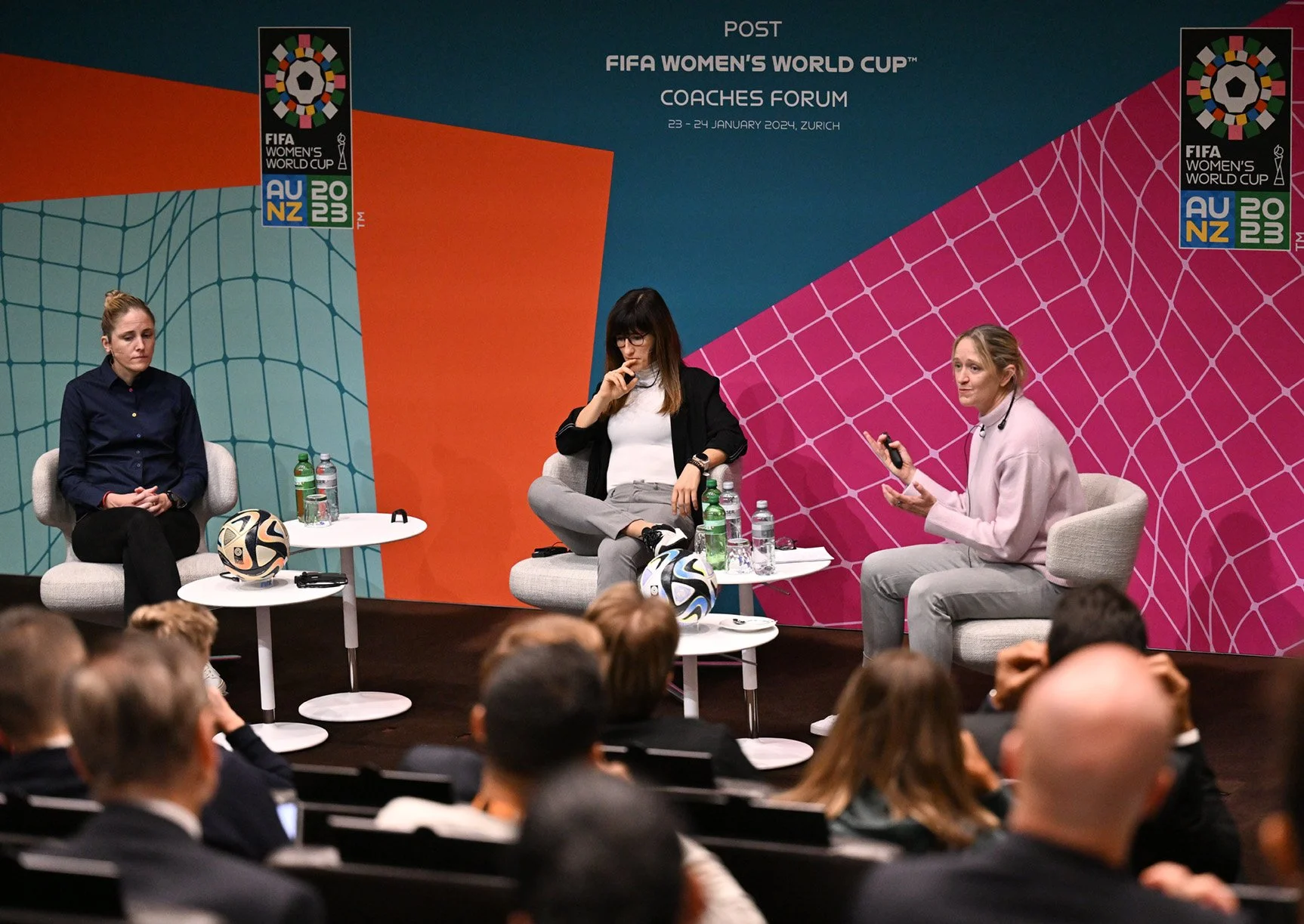 Three women seated on a stage at the FIFA Women's World Cup Coaches Forum, with an audience watching, and FIFA Women's World Cup branding in the background.