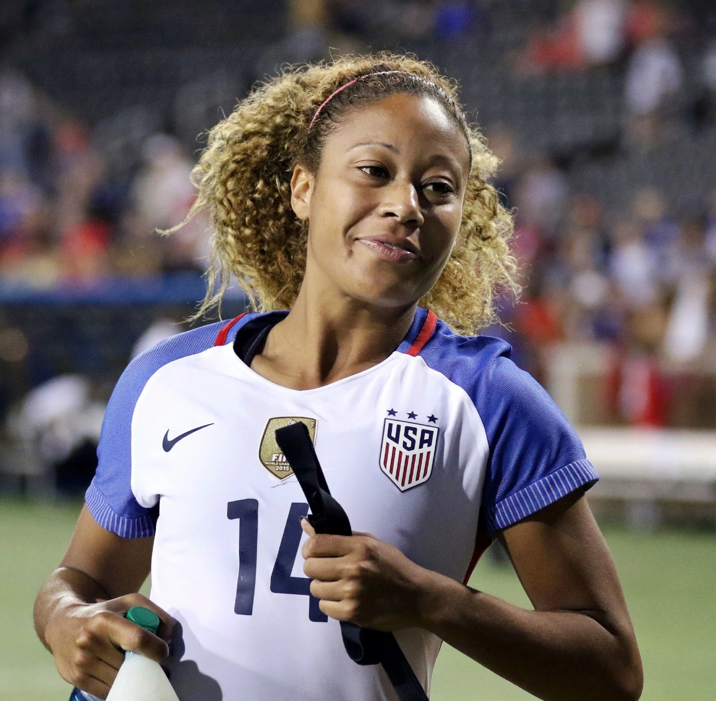 A female soccer player wearing a USA jersey with the number 14, holding a water bottle, standing on a soccer field with a blurred crowd in the background.