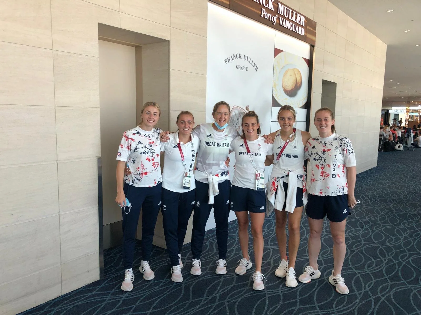 Six women posing together for a photo at an indoor venue, likely an airport, with some wearing uniforms and 0ne wearing a shirt with Great Britain printed on it, standing in front of a sign for Franck Muller Port of Vanguard.