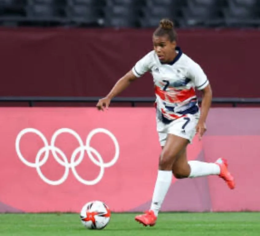 Female soccer player dribbling a ball on the field during the Olympics with Olympic rings in the background.