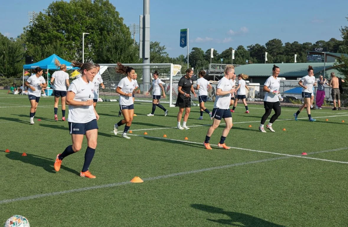 Soccer team warming up on field with cones during practice on a sunny day.