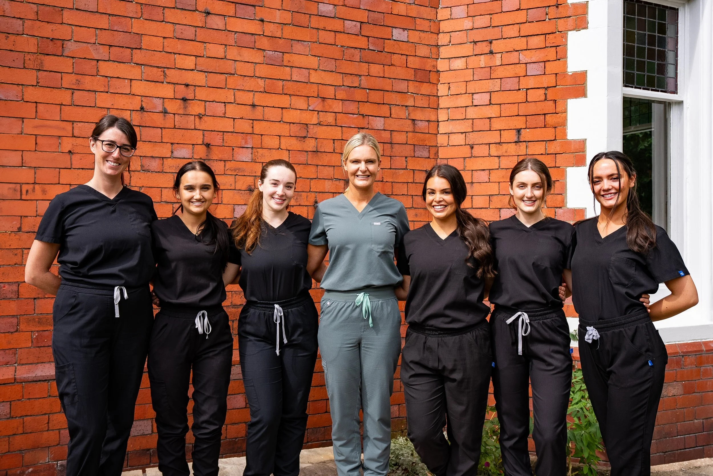 Group of seven diverse female healthcare professionals dressed in scrubs, standing outside against a brick wall, smiling at the camera.