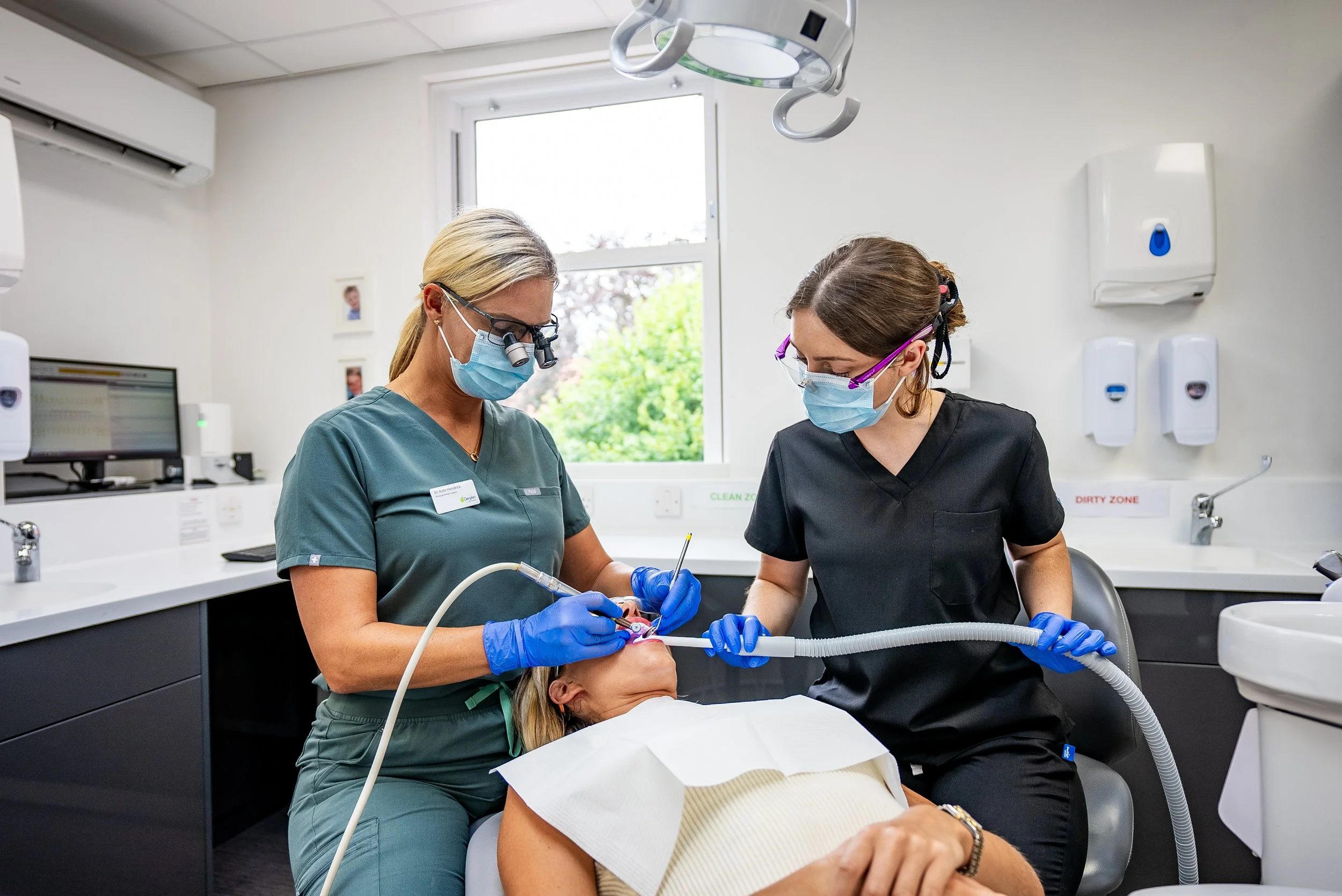 Two dental professionals working on a patient in a dental clinic, wearing masks and gloves, with dental equipment around them.