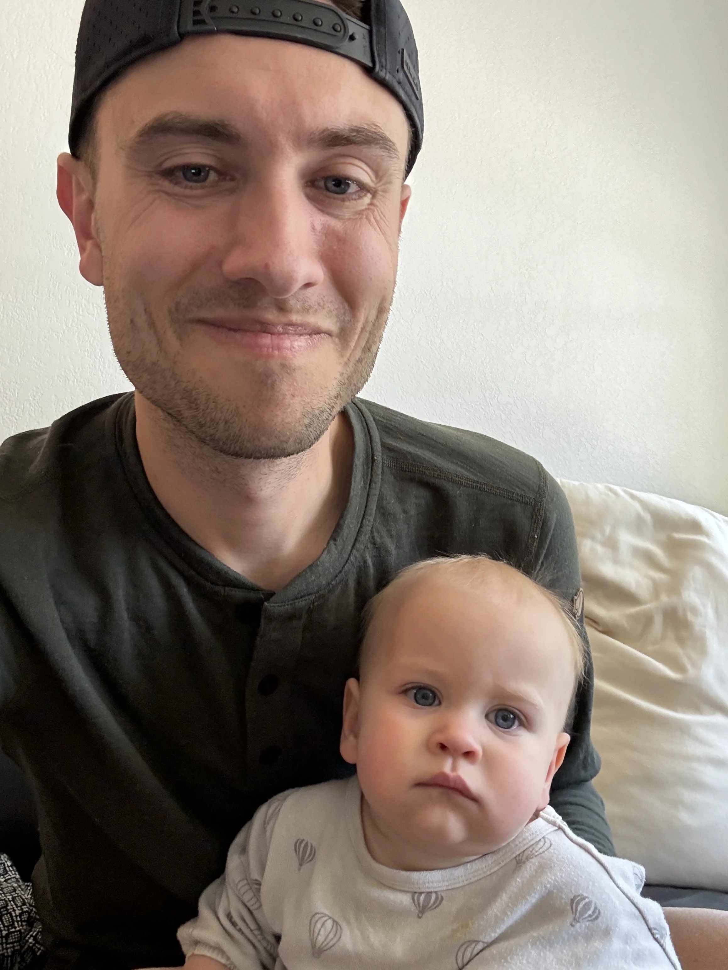 A man with short brown hair, wearing a black cap backwards, and a young child with light hair and blue eyes sitting together indoors against a white wall and a beige couch.