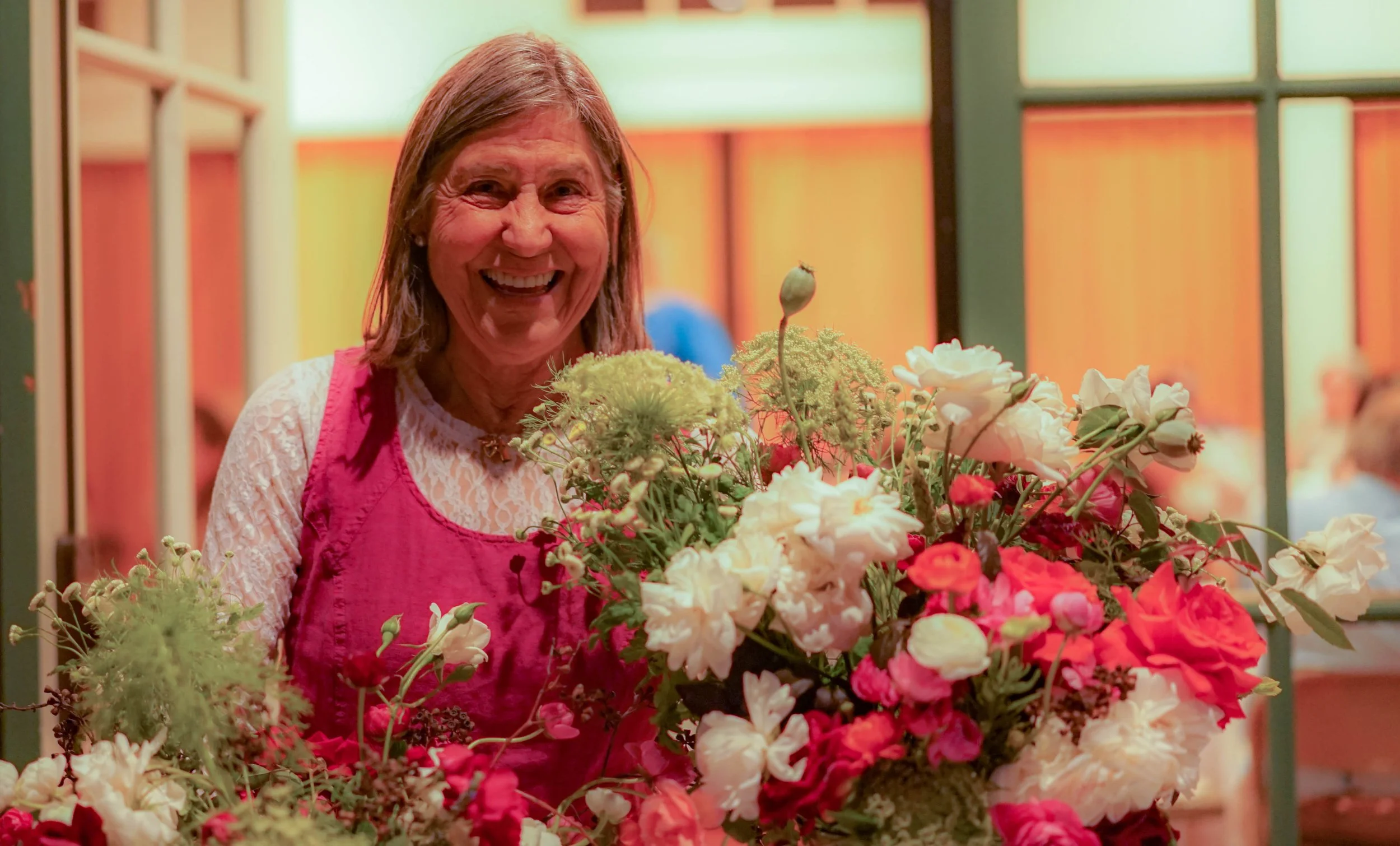 A woman with shoulder-length brown hair wearing a pink apron and a white lace top, smiling and standing behind a large bouquet of various colorful flowers including white, red, and pink blooms, inside a room with tan and green walls.