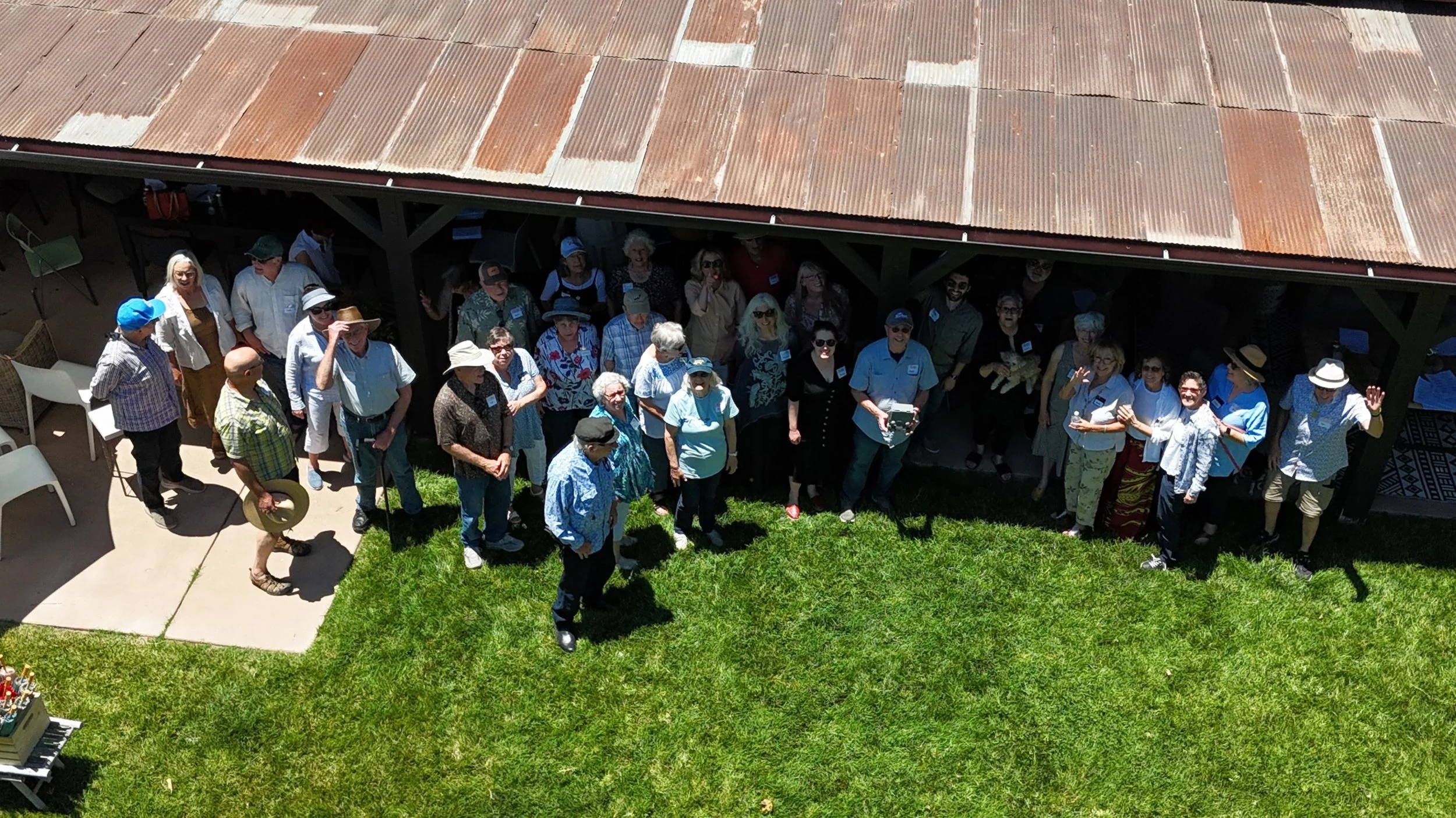 A group of people gathered outdoors on a grassy area, facing upward, possibly for a group photo or event, with a building with a tin roof in the background.