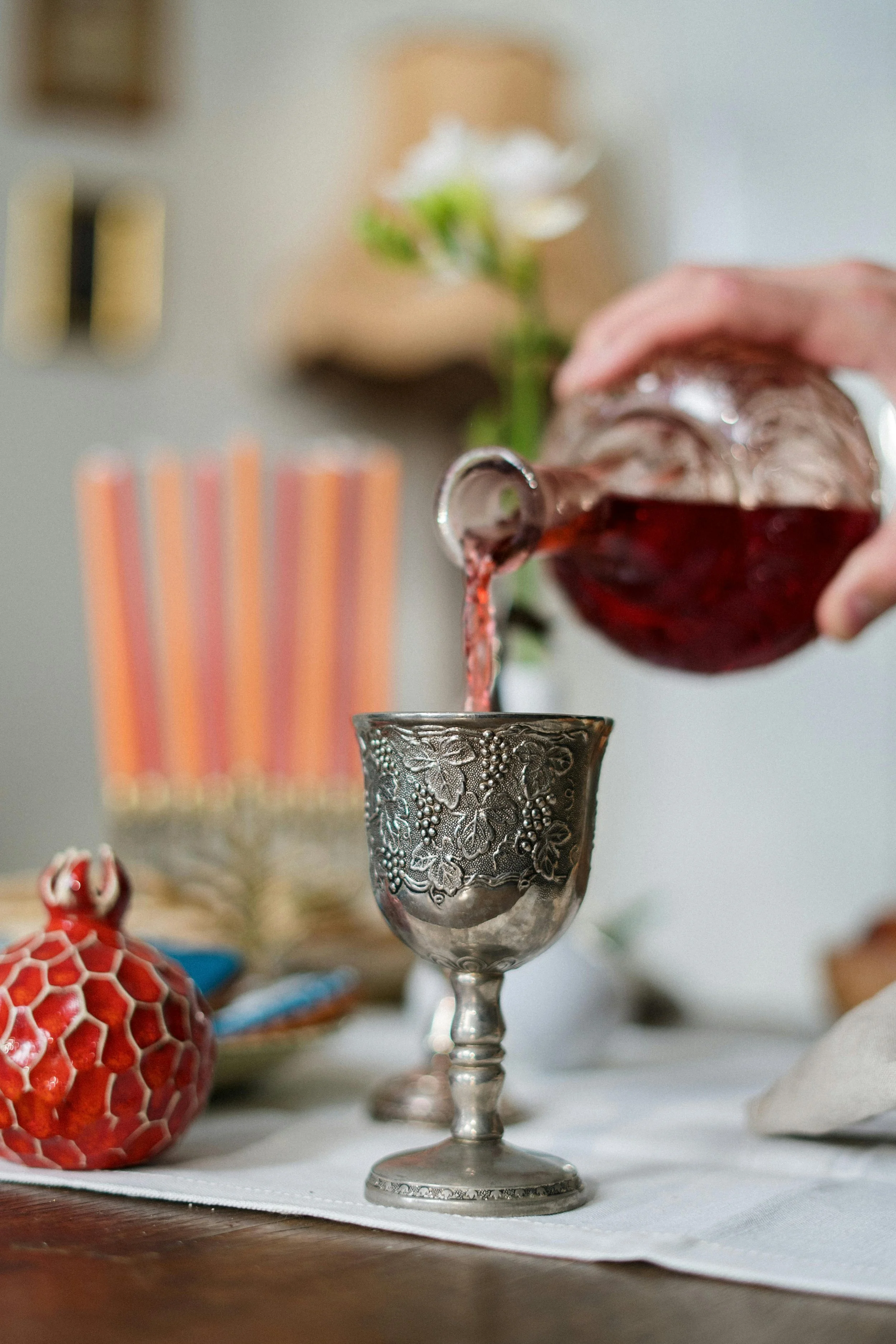 Person pouring red wine into a decorative silver wine glass on a dining table with a red ornament and birthday present in the background.