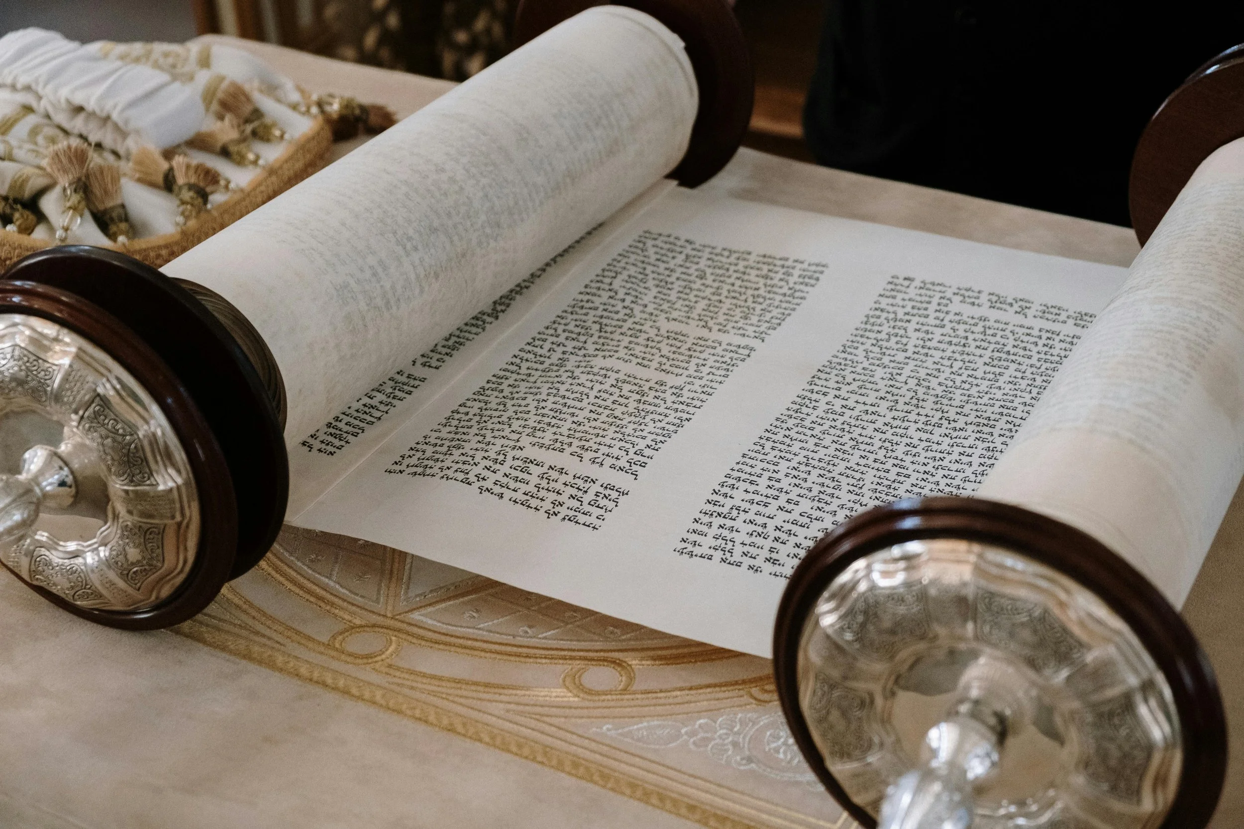 A Jewish Torah scroll partially unrolled on a table, with decorated silver handles and a cloth nearby.