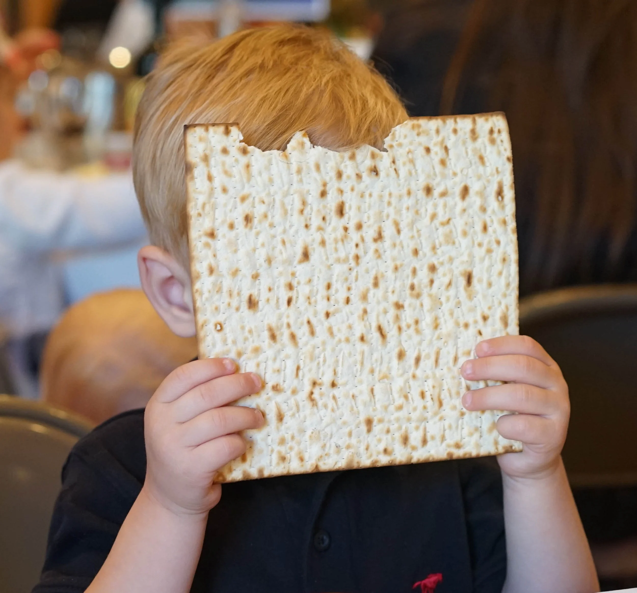 Child holding a large square piece of matzo covering their face, sitting in a dining area.