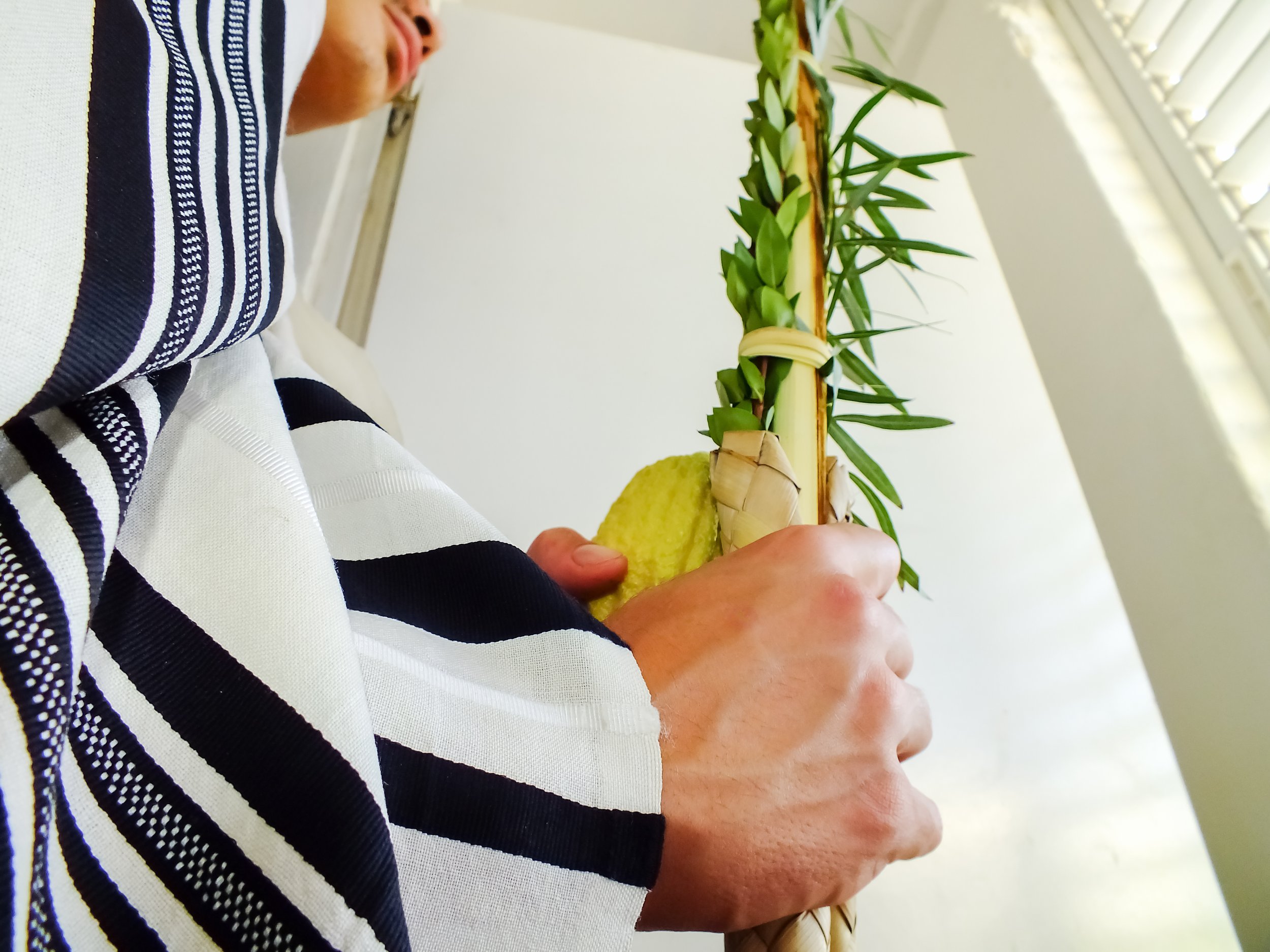 Person holding a palm leaf in a small woven basket, wrapped with green leaves, with a yellow object near their hand, indoors near window blinds.