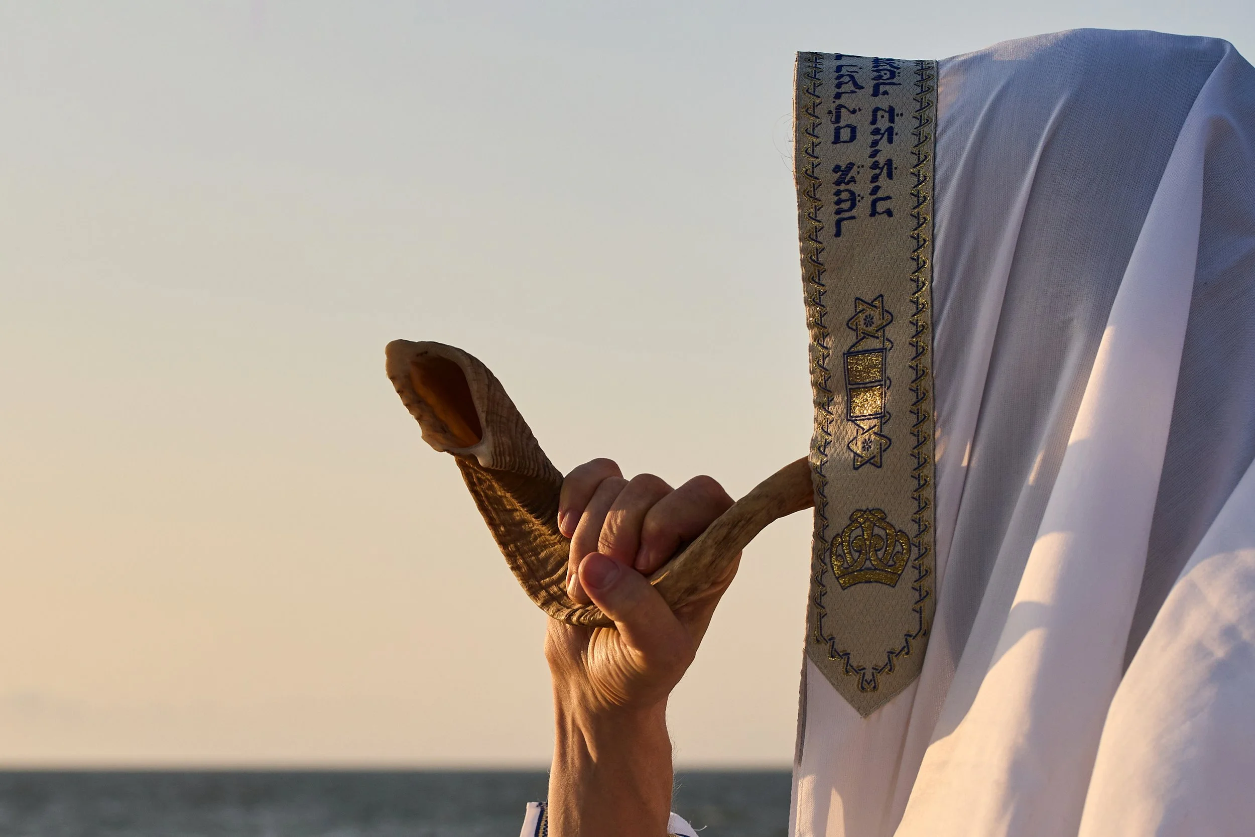 A hand holding a shofar, a ram's horn, near a Jewish prayer shawl with gold embroidery, outdoors at sunset.