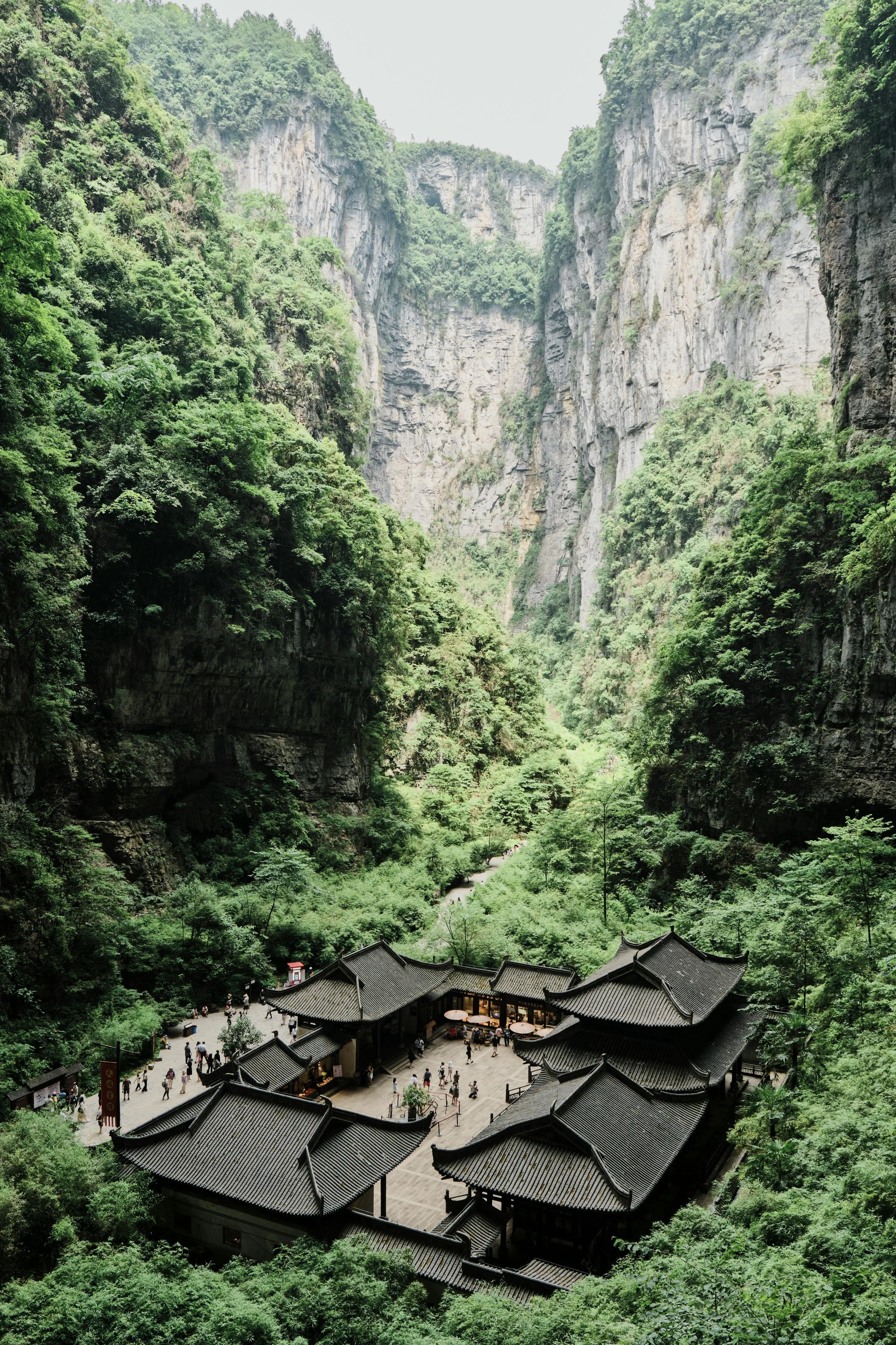 Ein Tempel in einem grünen Tal, umgeben von hohen Klippen mit dichtem Wald. Es sind viele Menschen auf dem Tempelgelände und im umliegenden Bereich unterwegs.