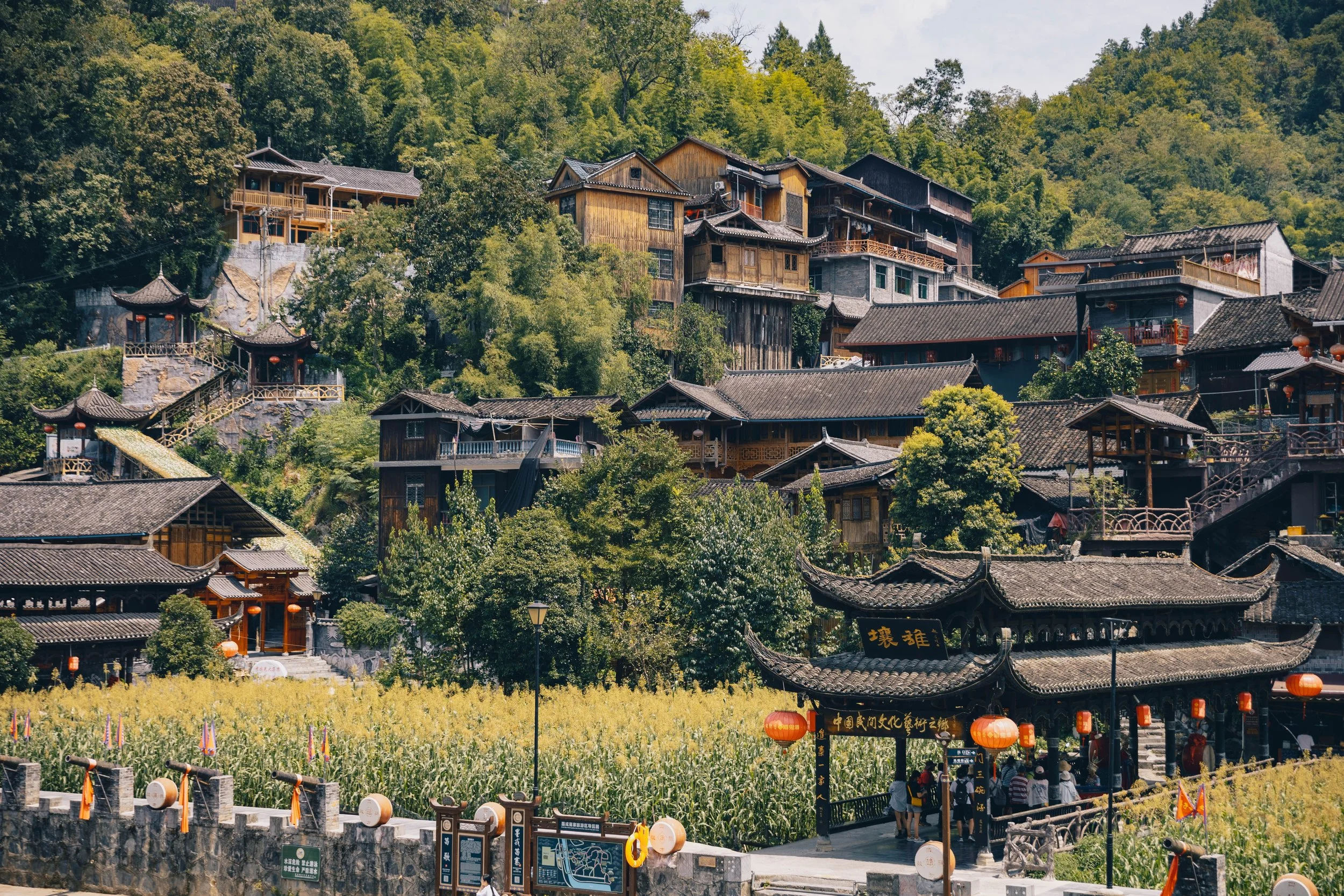 Traditionelle chinesische Architektur aus der Region Guizhou von ethnischen Einheimischen Miao auf einem Hügel, umgeben von grünen Bäumen, mit Pagoden, Holzhäusern und roten Laternen im Vordergrund.