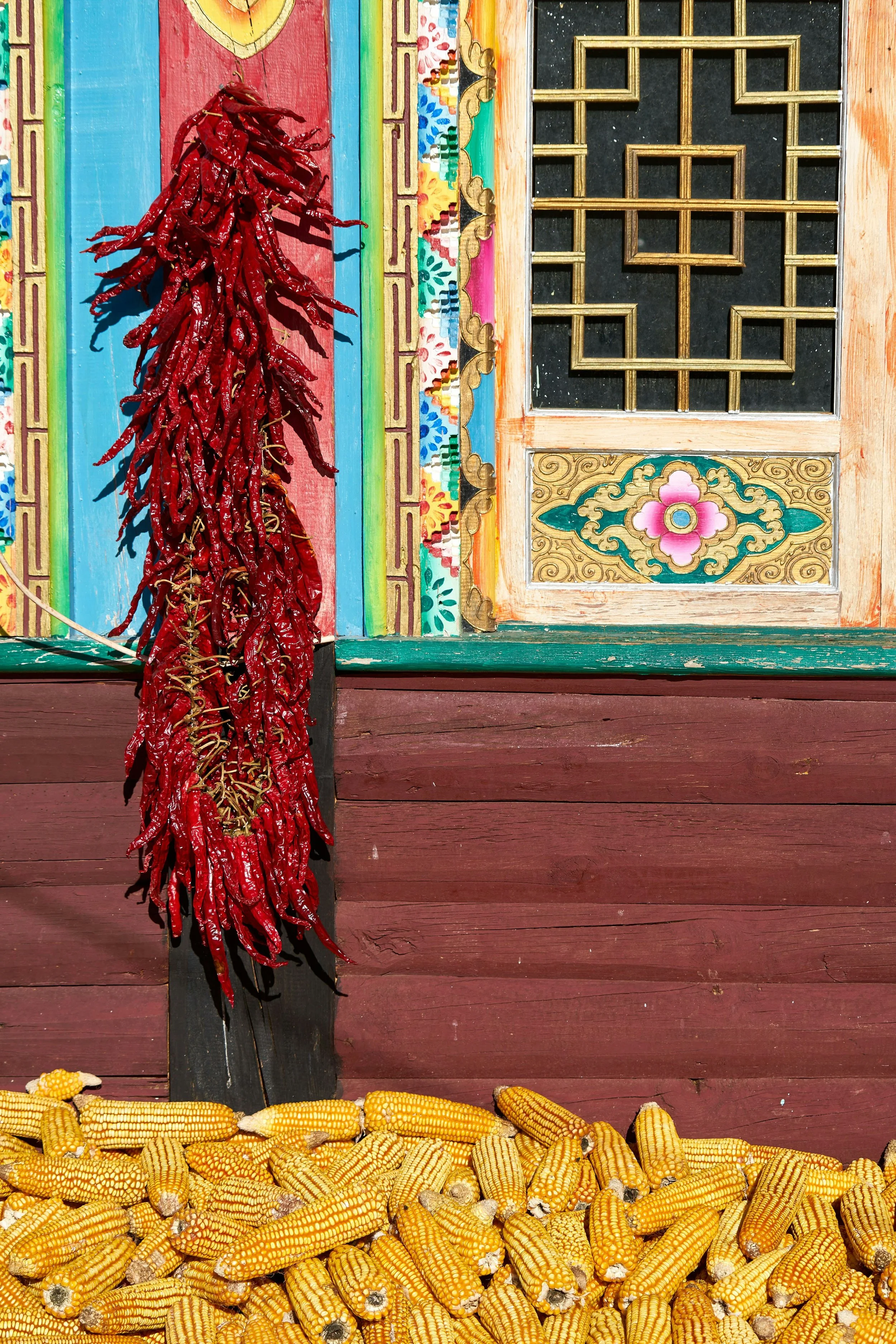 Bündel rote Chilischoten hängen an einer schwarzen Holzkufe vor einem bunten, kunstvoll asiatisch verzierten Fensterrahmen. Darunter liegen viele gelbe Maiskolben auf einer Holzoberfläche. Eine Szene aus einem asiatischen Dorf.