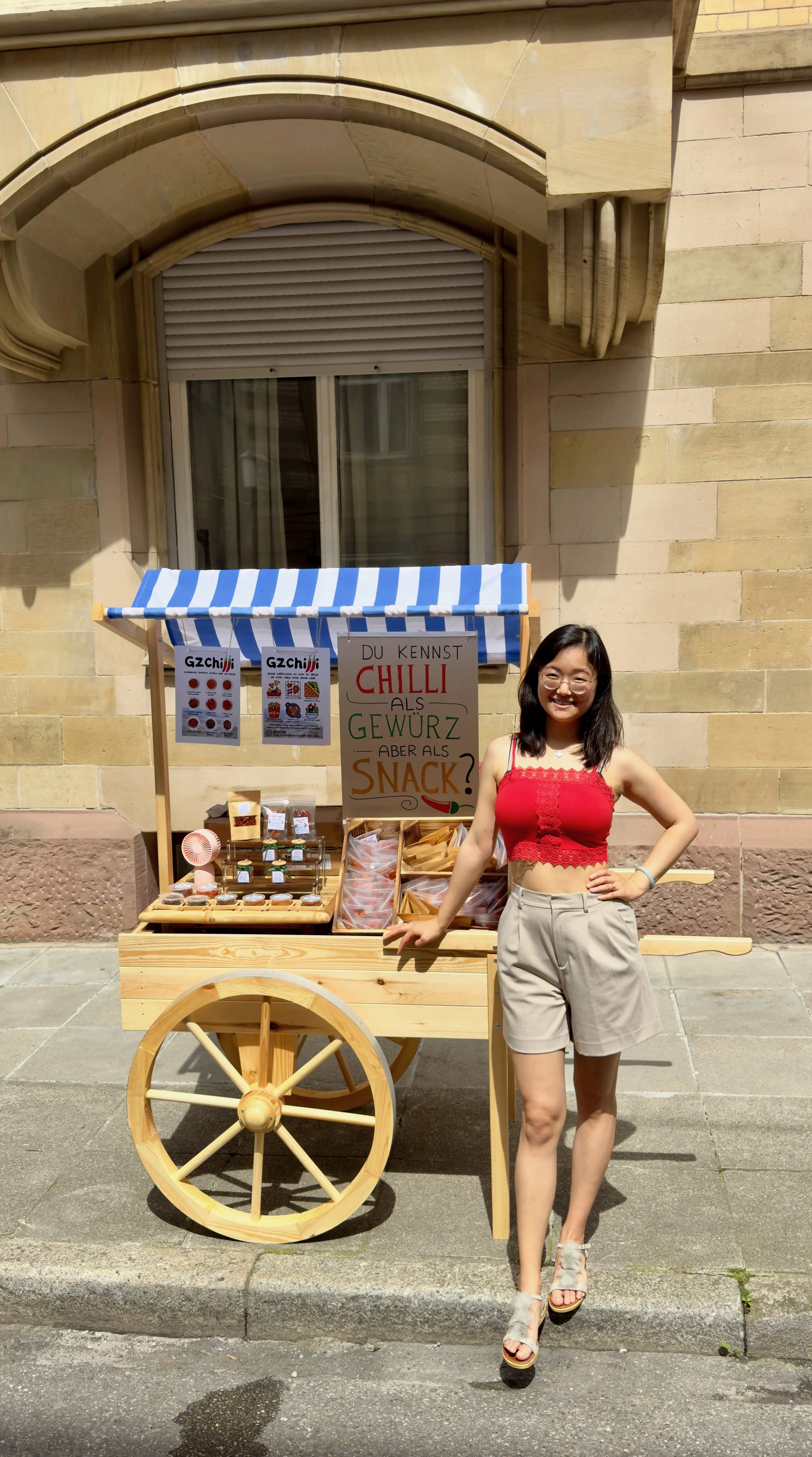 Eine junge Frau steht vor einem Holzwagen, der Gewürze verkauft. Das Verkaufsstand hat ein blau-weiß gestreiftes Dach und Schilder mit der Aufschrift: "Du kennst Chilli als Gewürz, aber als Snack?" auf Deutsch. Die Frau trägt eine rote ärmellose Top und bequeme Shorts, hat lange dunkle Haare und trägt Brille. Sie lächelt und posiert vor dem Stand, der in einer Stadt mit Backsteinmauern im Hintergrund steht.