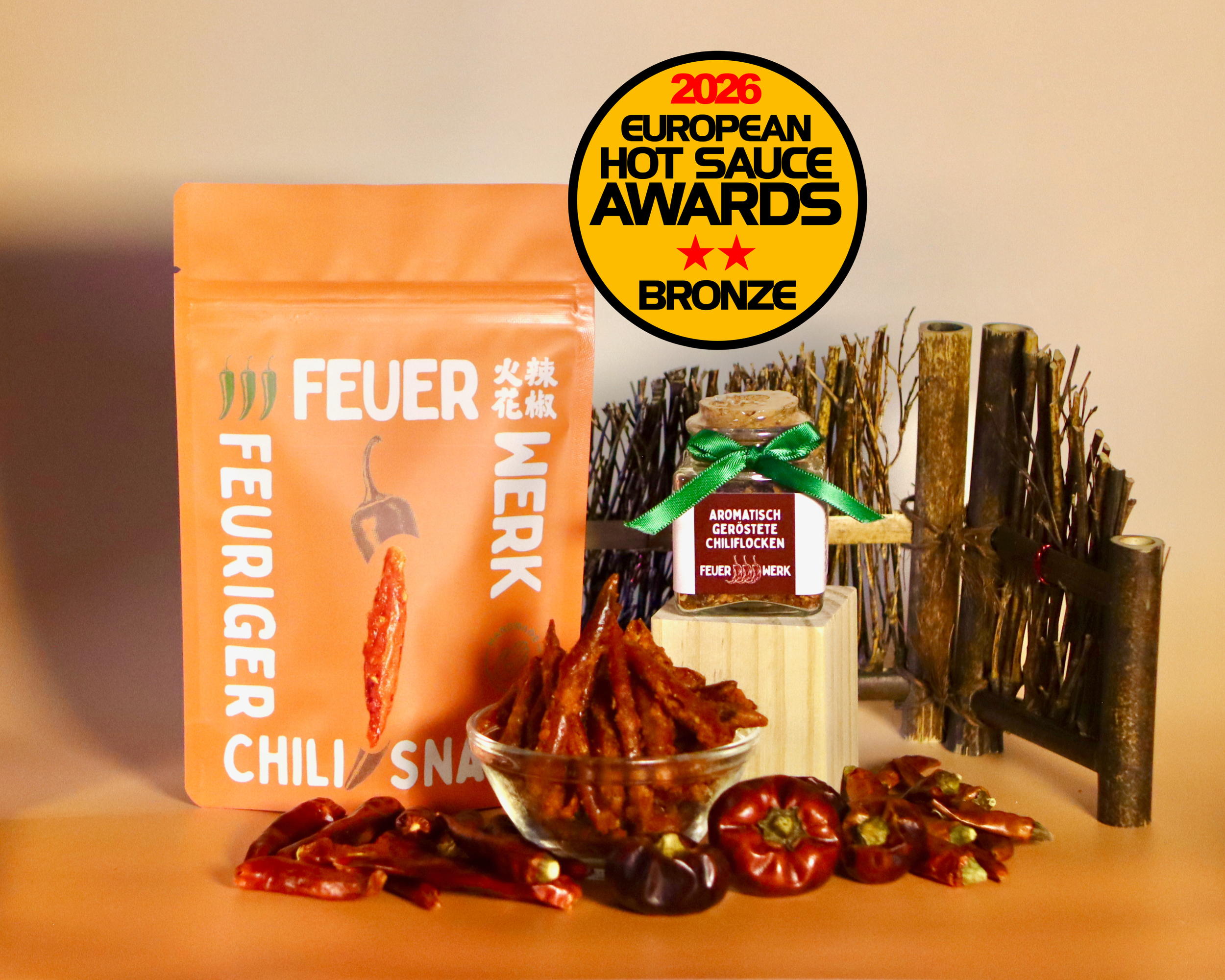 Ein Glas mit getrockneten Chilischoten, umgeben von mehr Chilischoten, hinter einem orangefarbenen Paket mit chinesischen und deutschen Schriftzeichen und einem Logo, das den European Hot Sauce Award 2026 Bronze-Auszeichnung zeigt.