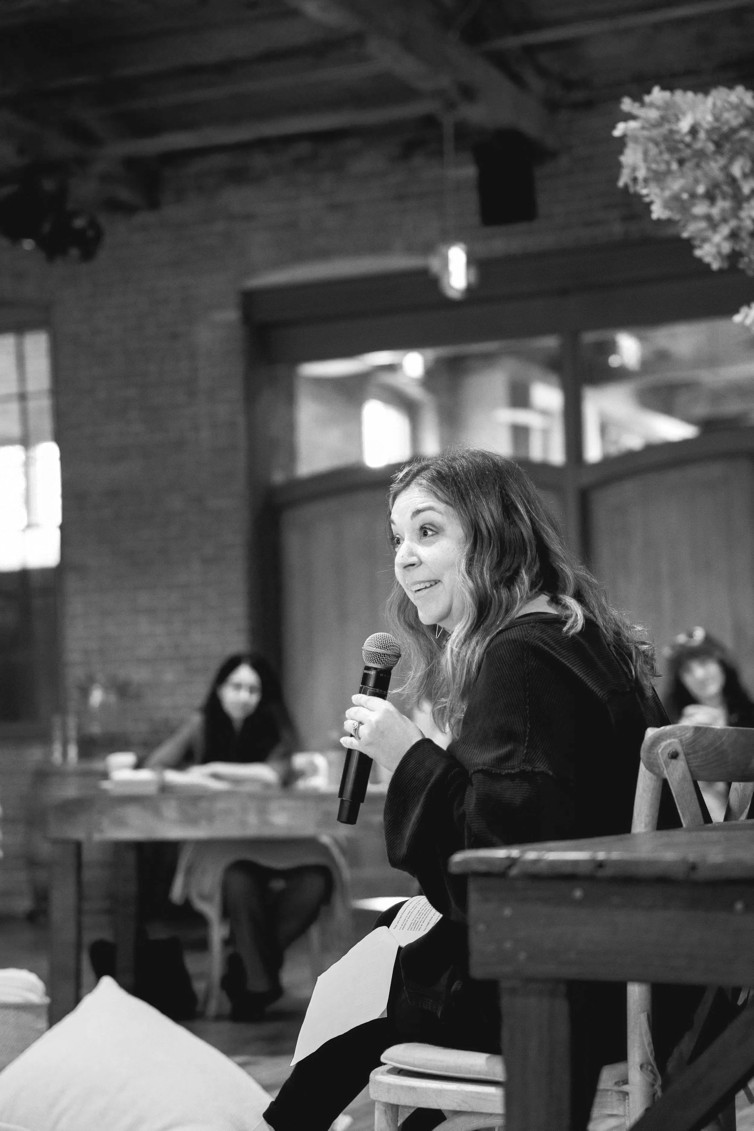 A woman, Andi Campbell, with shoulder-length wavy hair holding a microphone and speaking during a discussion or presentation in an indoor setting with other seated women in the background.