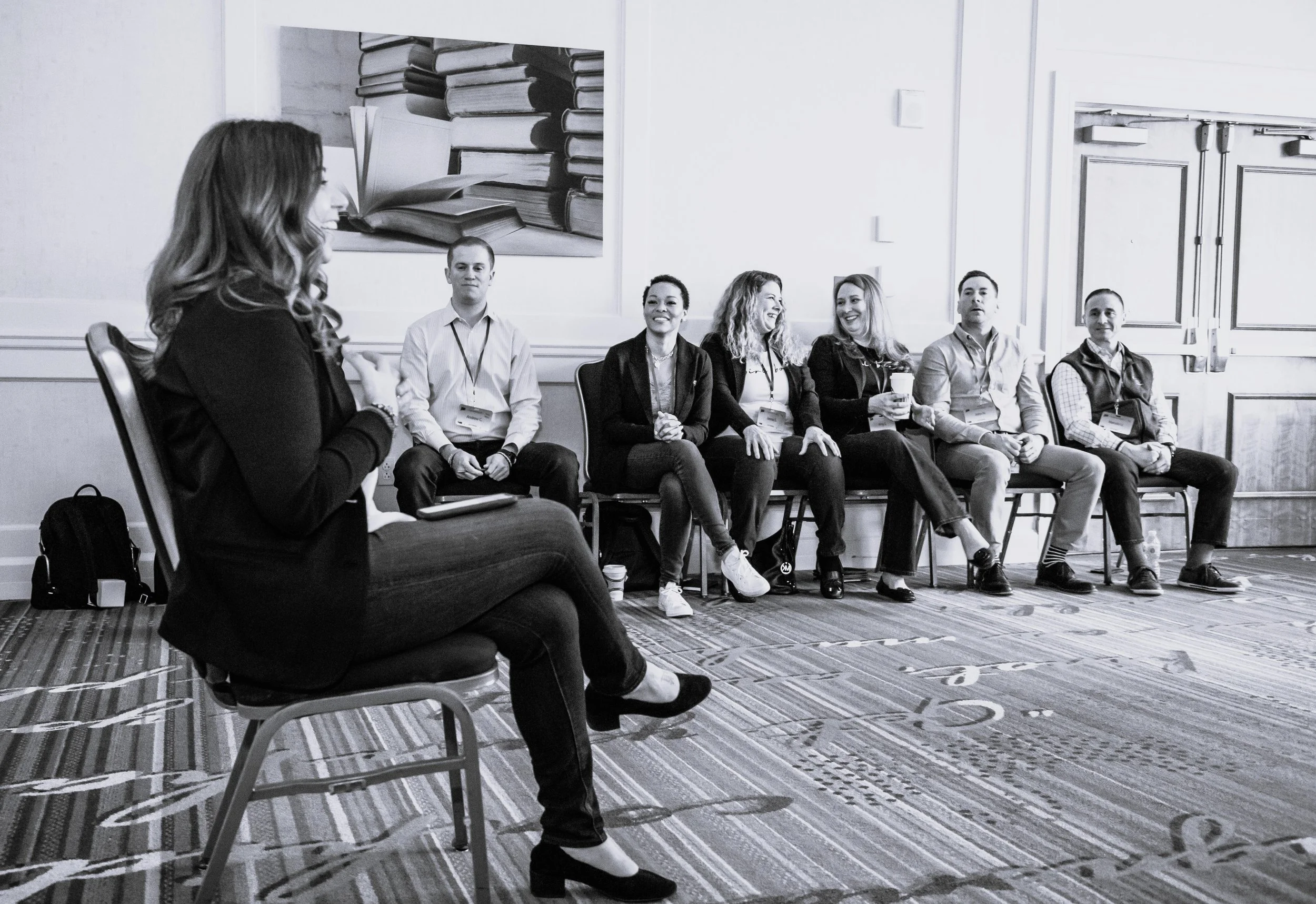 A woman, Andi Campbell, speaking at a panel or workshop, seated on a chair, with seven people sitting in a row listening to her, in a conference room with artwork of stacked books on the wall.