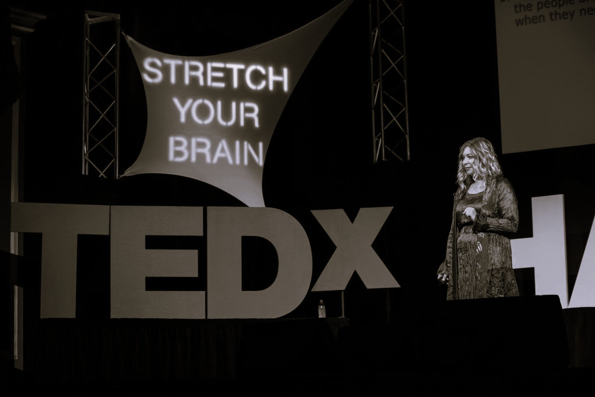 A woman, Andi Campbell, with curly hair on stage at a TEDx event, speaking in front of large TEDx letters and a screen projecting the message 'Stretch Your Brain'.