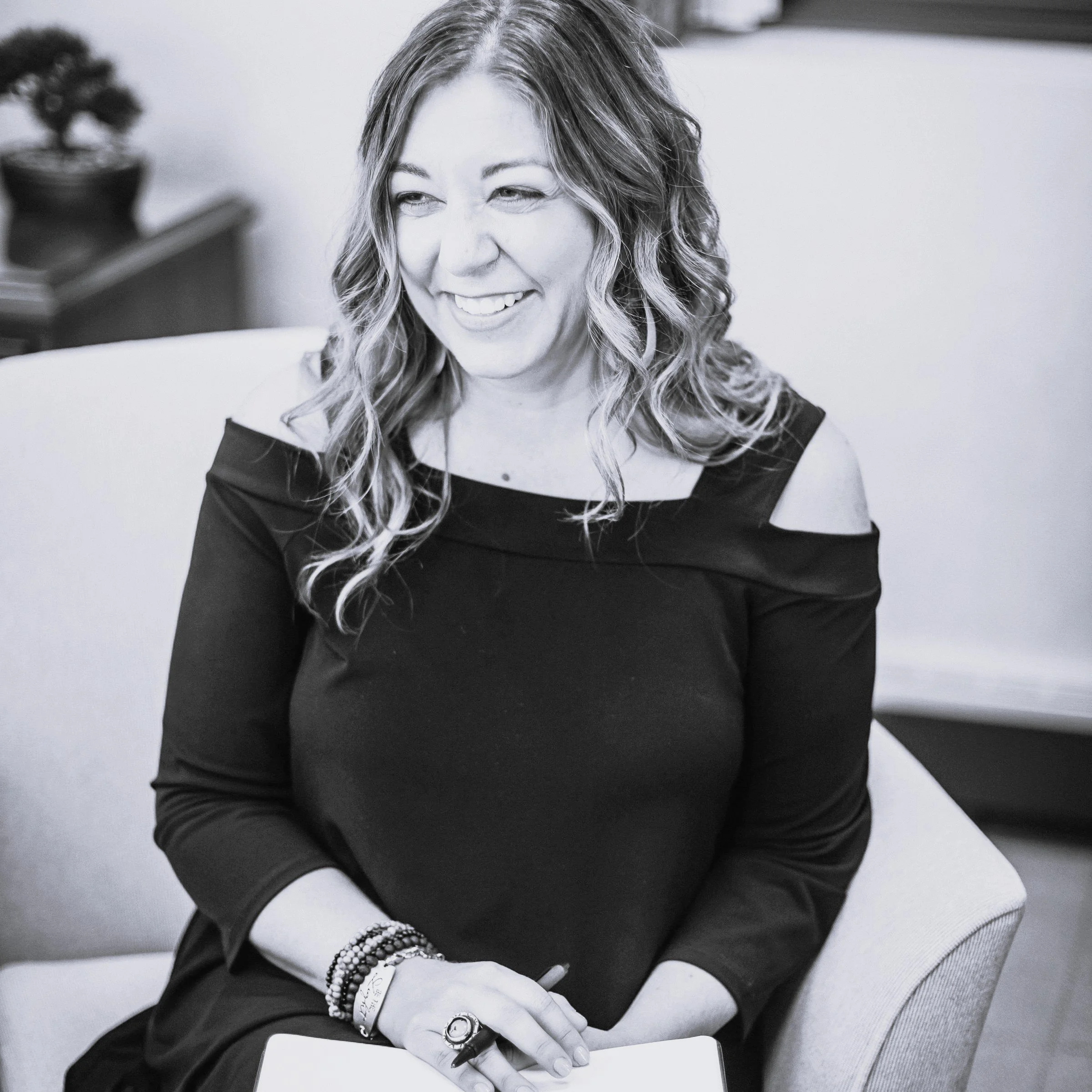 Andi Campbell, a woman, with wavy hair smiling and sitting in a chair, wearing a black cold-shoulder top and jewelry.