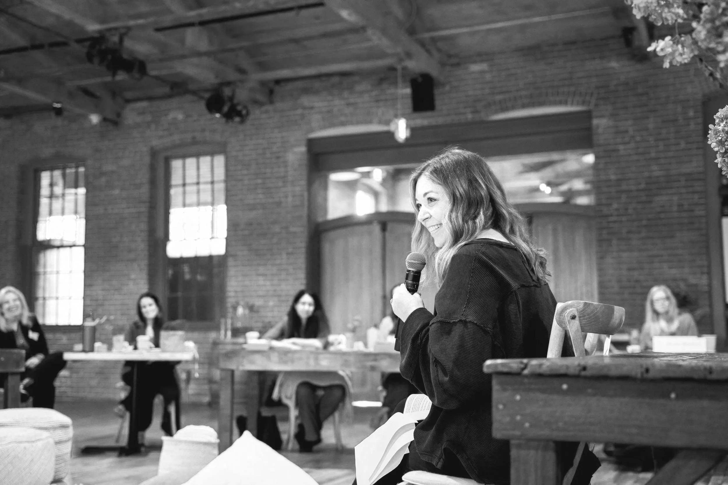Andi Campbell, a woman, sitting on a chair and speaking into a microphone at a panel discussion. Four women are seated at tables in the background in a room with brick walls and large windows, with the image in black and white.