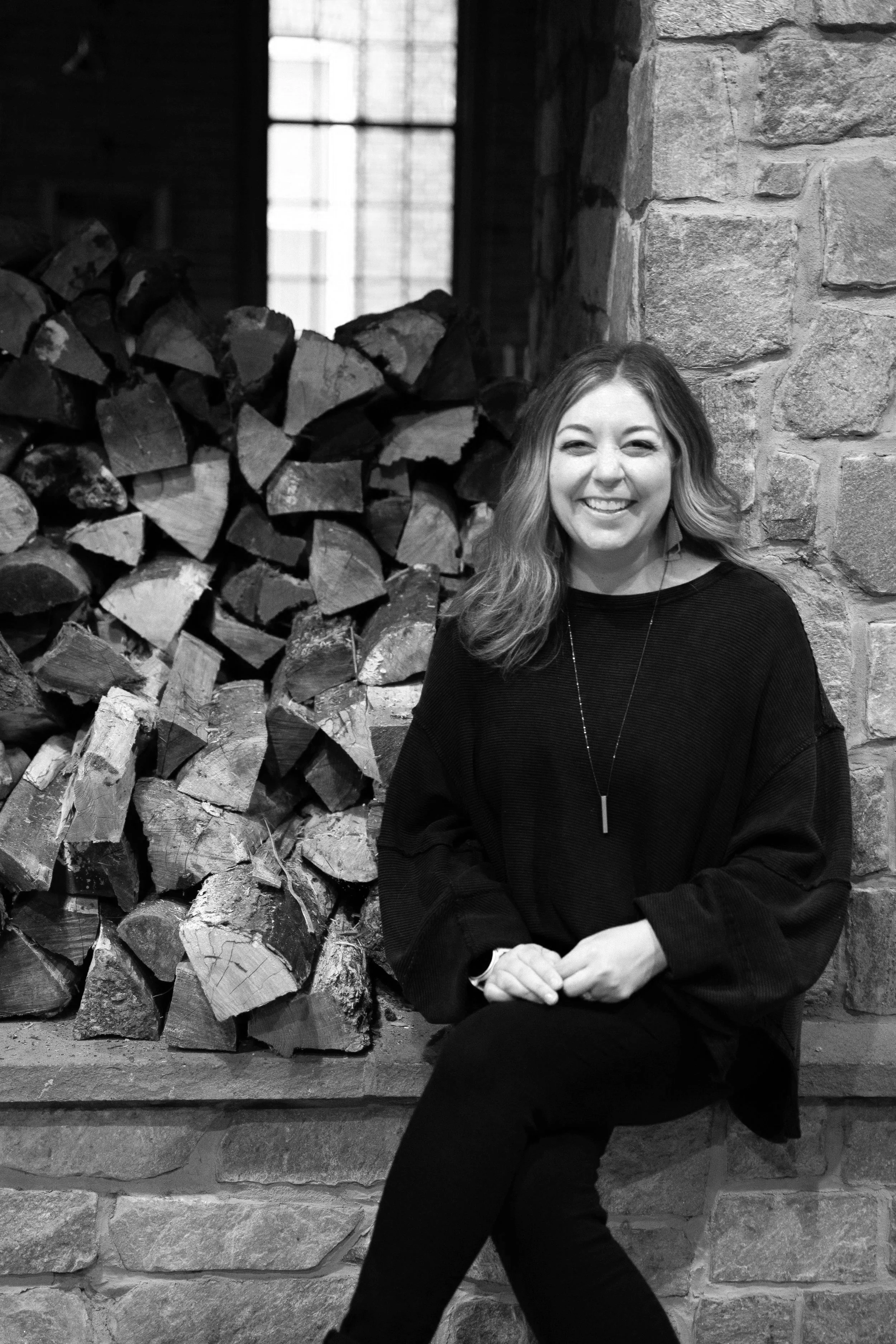 Andi Campbell, a woman, with wavy hair smiling, sitting in front of a stacked pile of firewood inside a rustic room with a stone wall and a window.