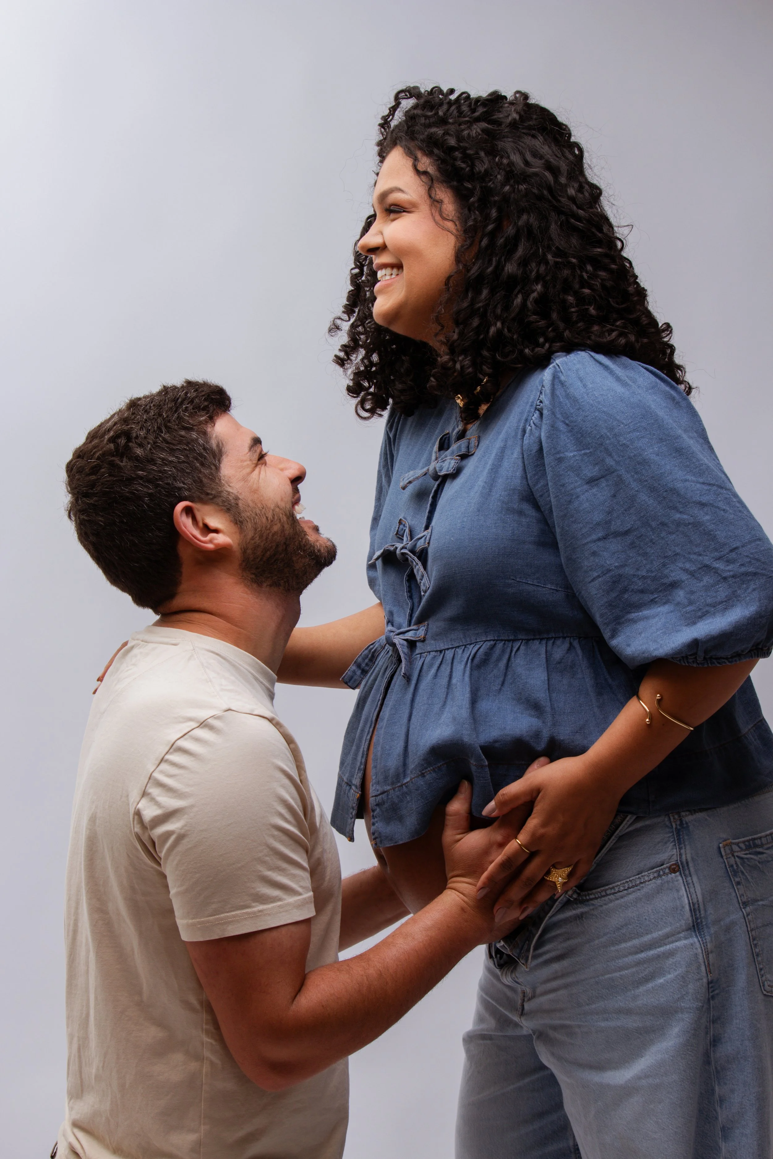 A man and woman are smiling and looking at each other, with the man kneeling and holding the woman's pregnant belly.