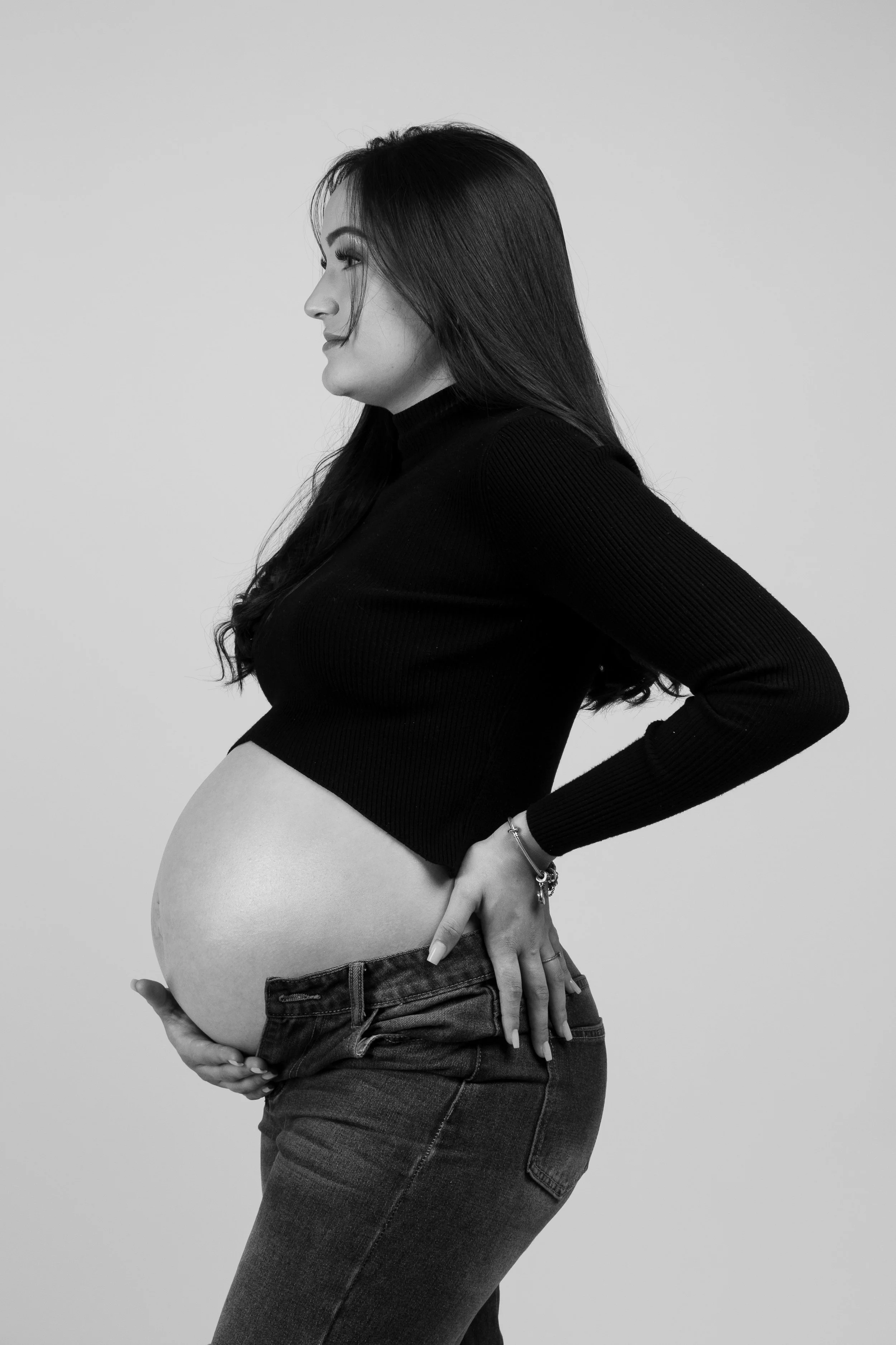 Black and white photo of a pregnant woman in profile, wearing a black long-sleeve top and jeans, with her hand resting on her belly.